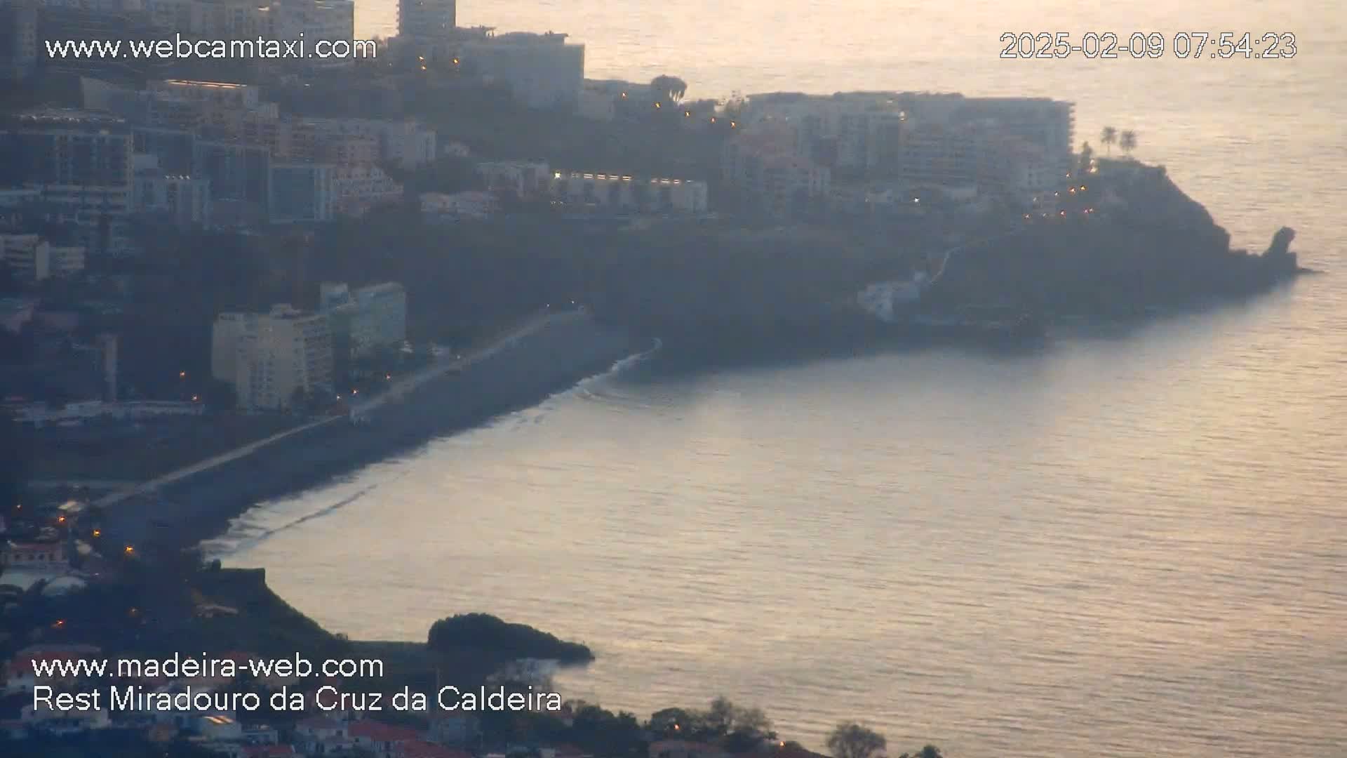 A coastal city at sunrise with calm waters lapping a dark-sand beach next to a cliffside.