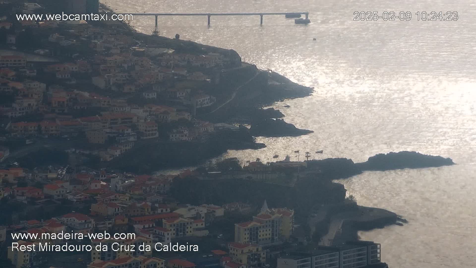 A coastal town nestled on a rocky hillside overlooking a calm, sunlit ocean with a distant bridge visible.