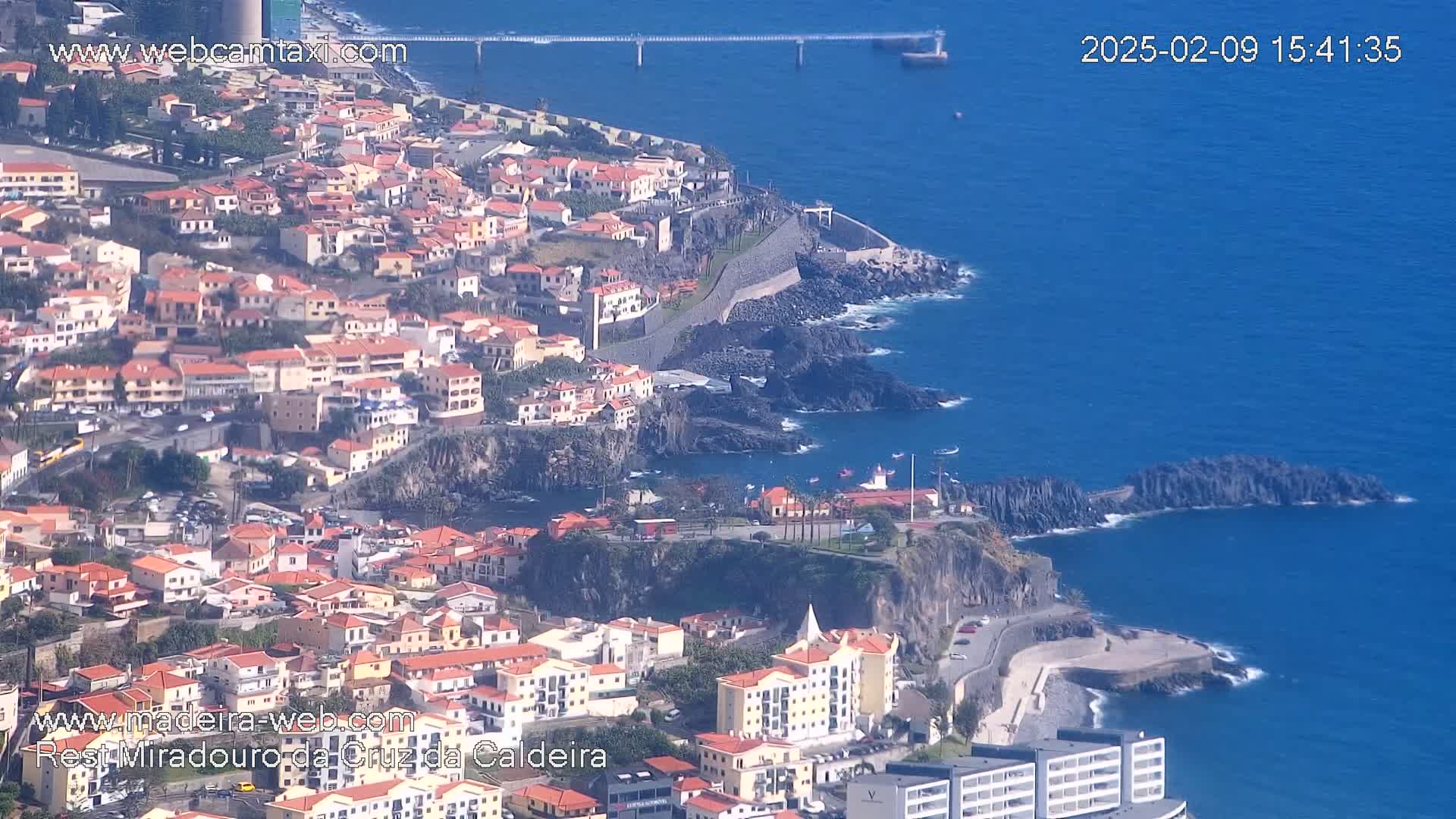 An aerial view shows a coastal town with red-roofed buildings clustered on a hillside, overlooking a dark blue ocean on a sunny day.