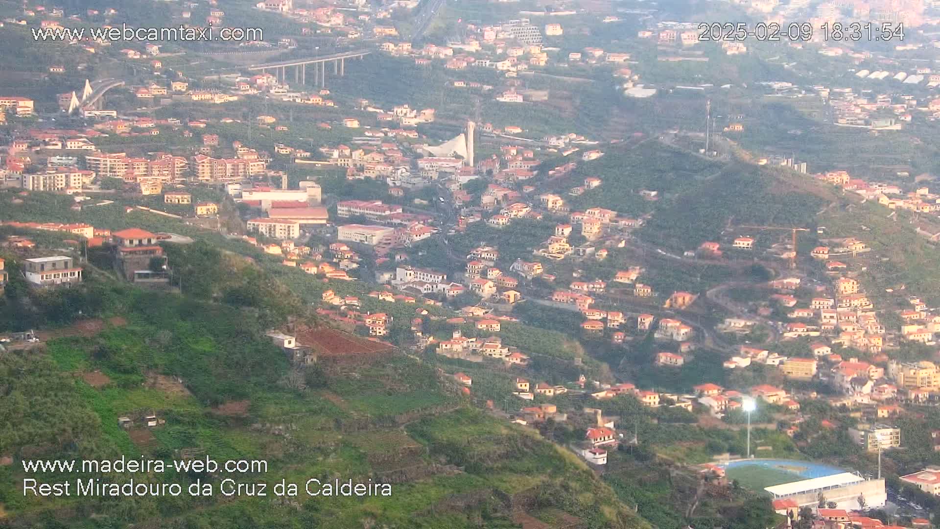 A hazy, high-angle view shows a hillside town with numerous low-rise buildings, terraced farmland, and a distant bridge.