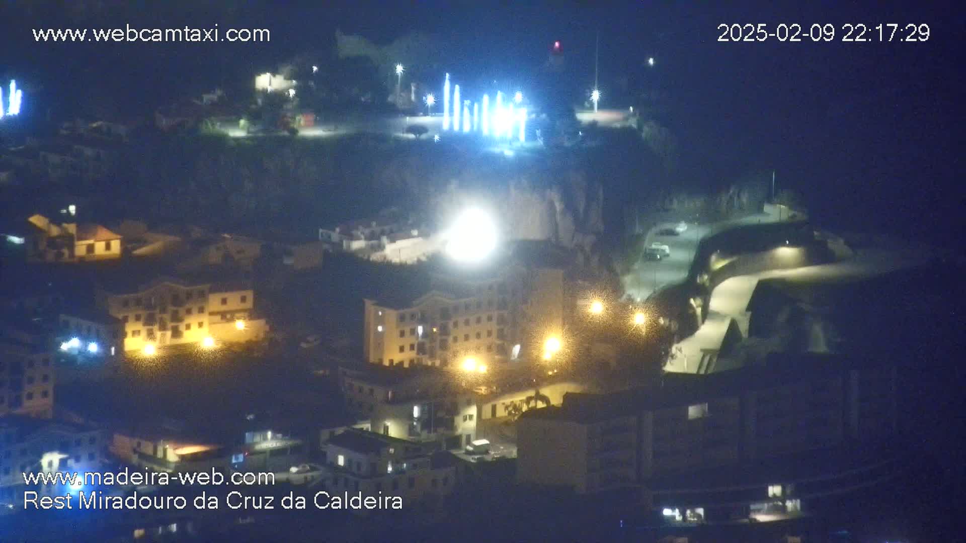 A nighttime, high-angle view shows a town illuminated by streetlights and building lights, with a bright light source in the center and a series of vertical lights on a hill in the background.