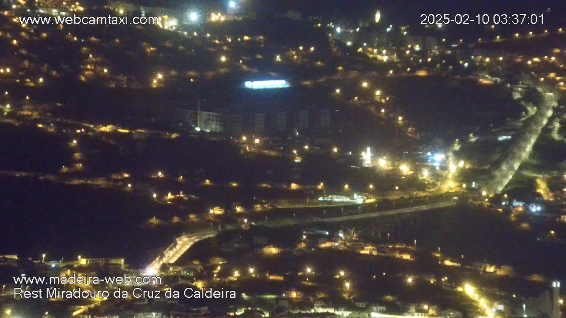 A nighttime aerial view of a hilly town illuminated by numerous lights.