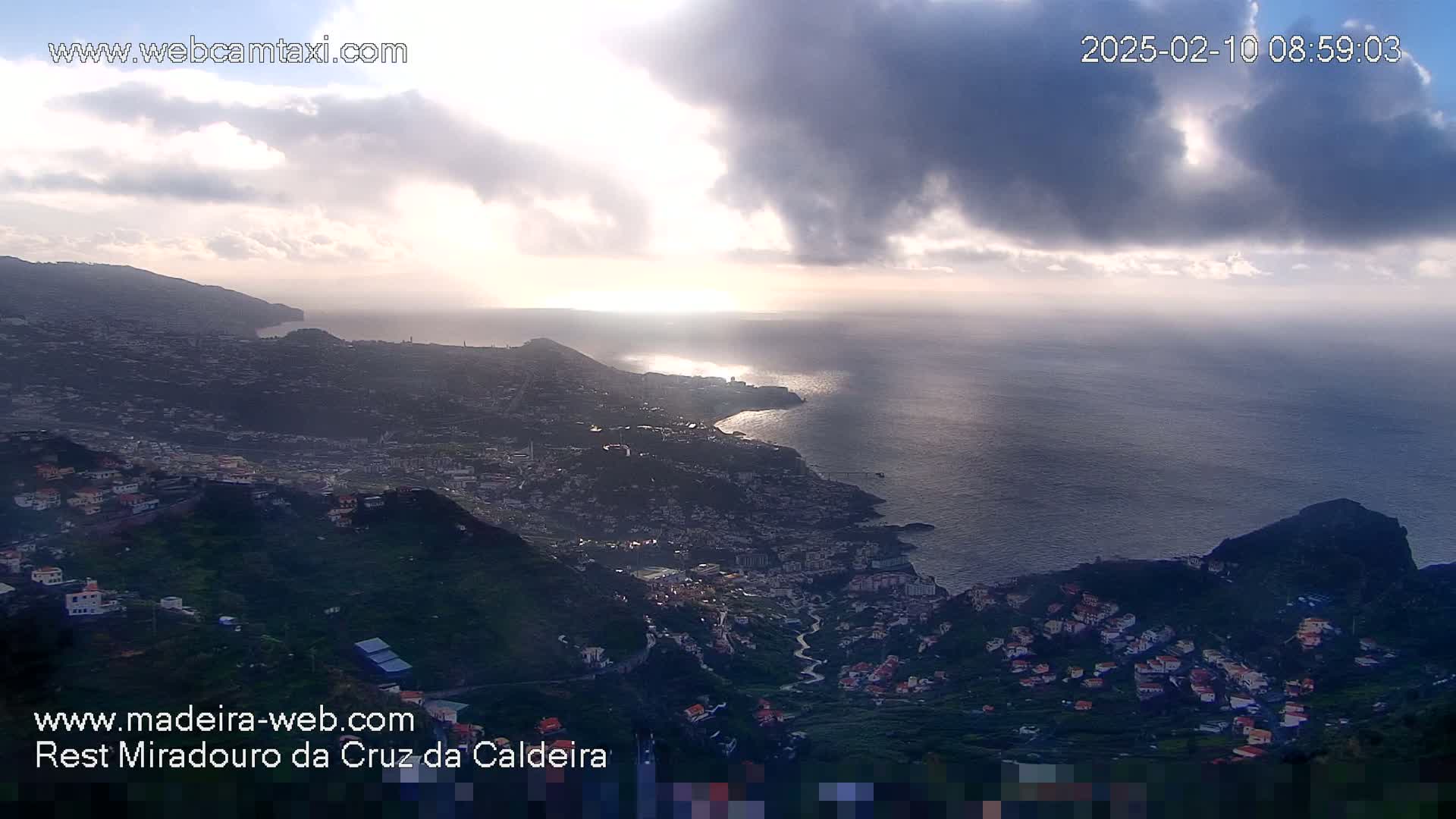 A coastal town nestled in hillsides overlooks a calm ocean under a partly cloudy sky, with the sun shining through the clouds.