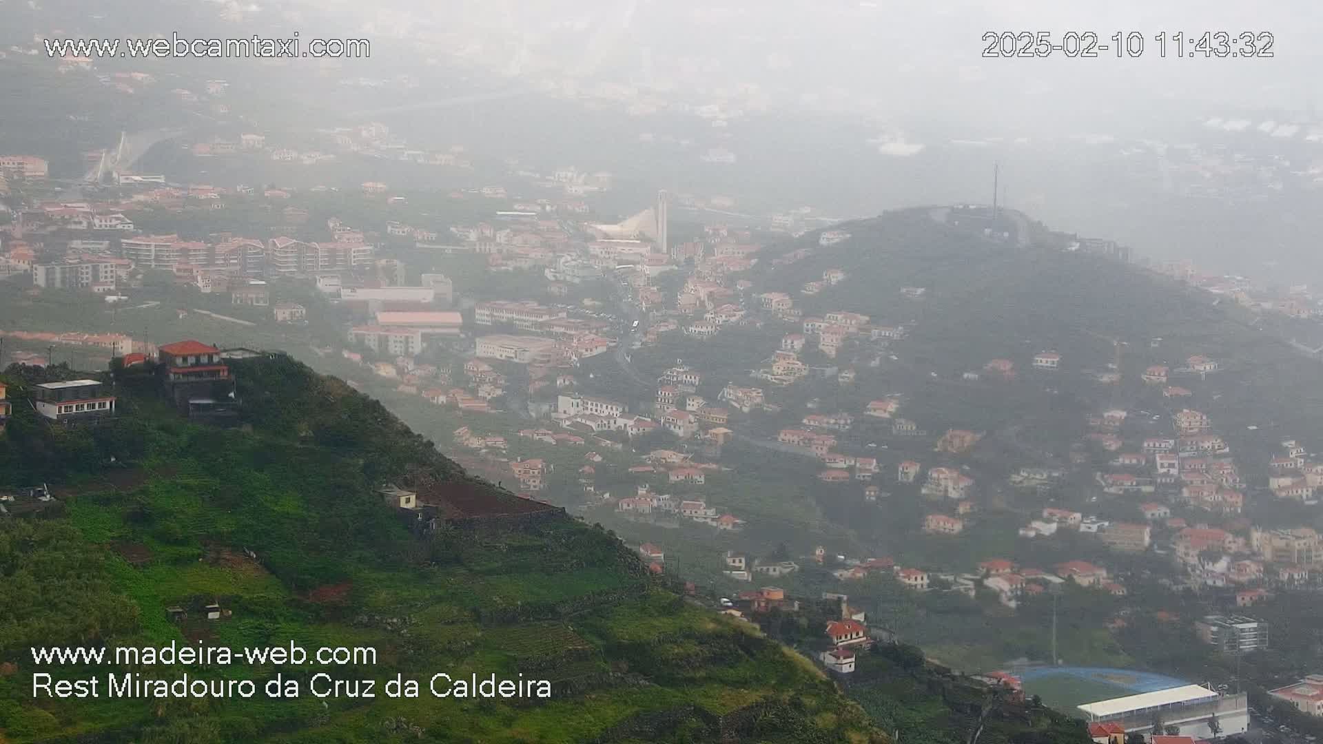 A hazy, rain-swept view shows a hillside town built on steep slopes, with buildings cascading down the mountainside.