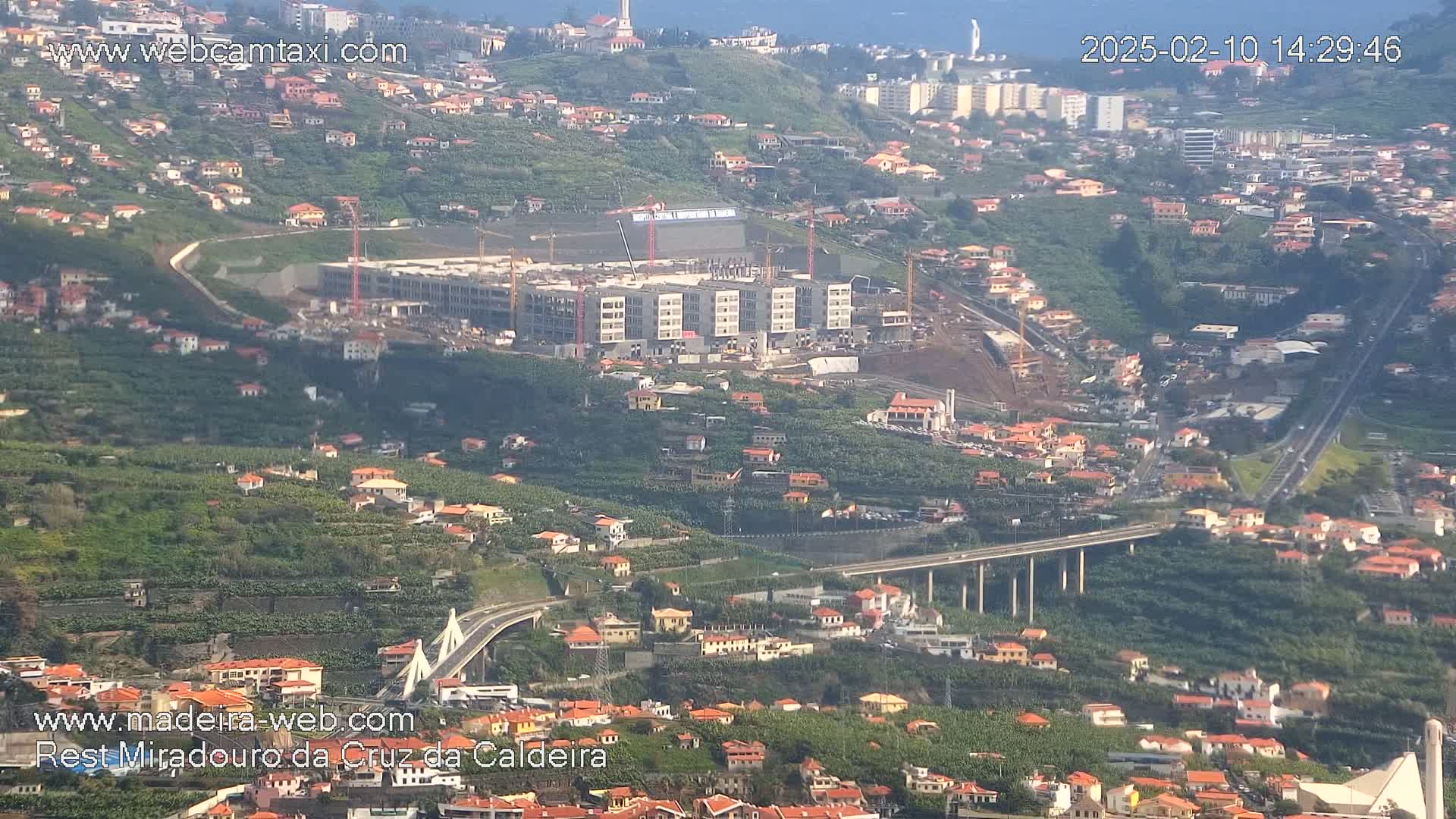 A large building under construction is situated on a hillside overlooking a highway and bridge, with numerous houses and lush greenery covering the landscape on a clear day.