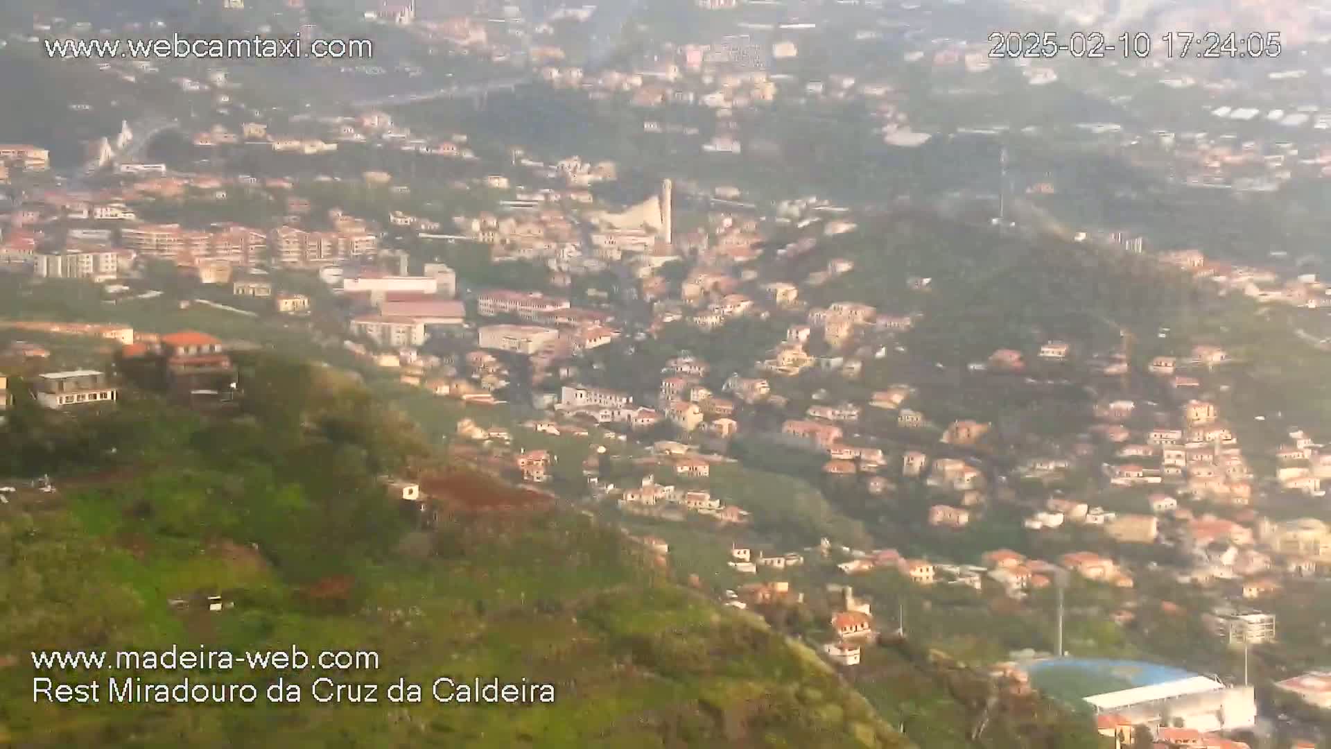 A hazy, long-distance view shows a hillside town with many buildings nestled among green vegetation.