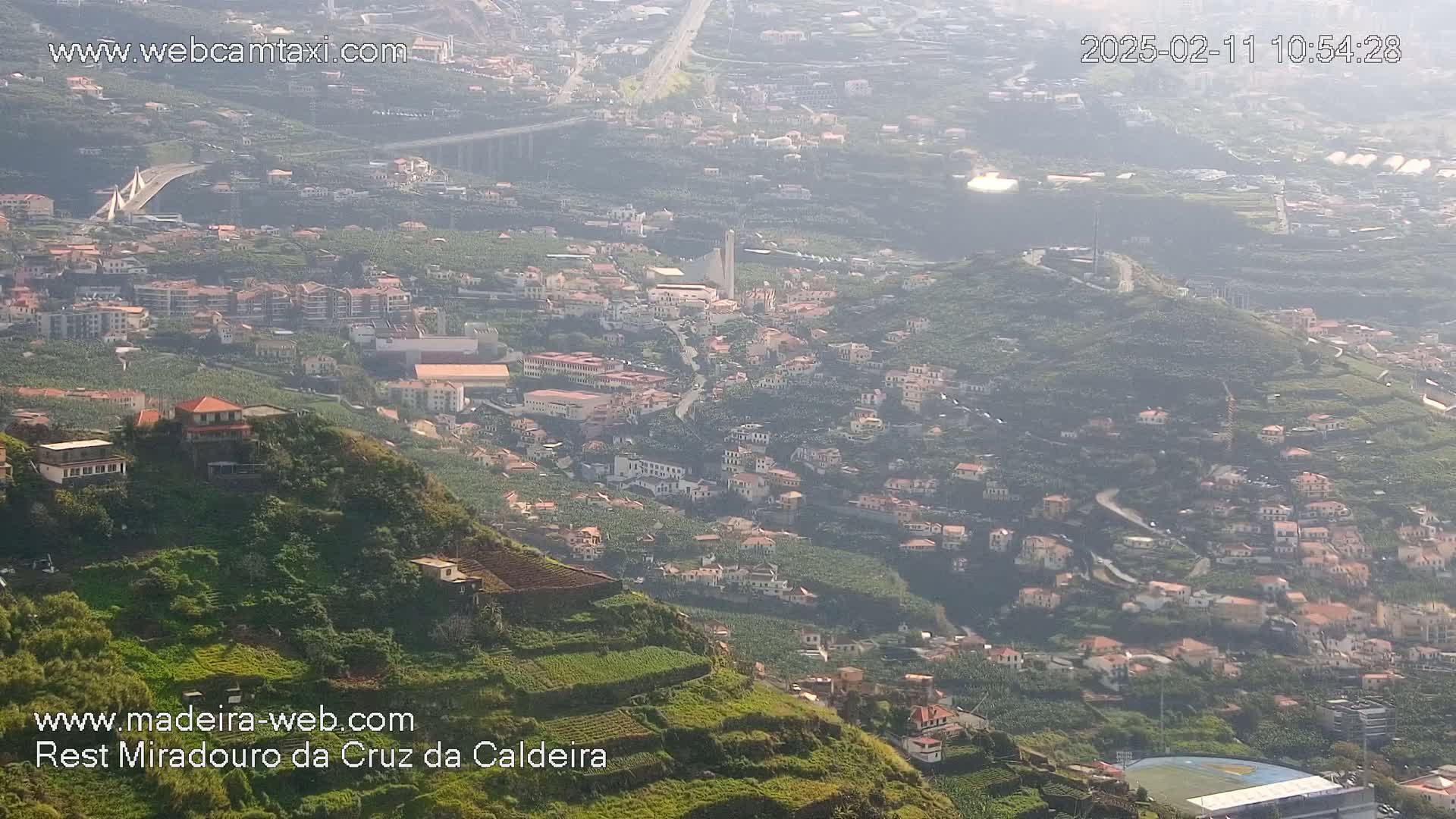 A hazy view shows a hillside town nestled amongst terraced farmland, with a bridge and highway visible in the distance.