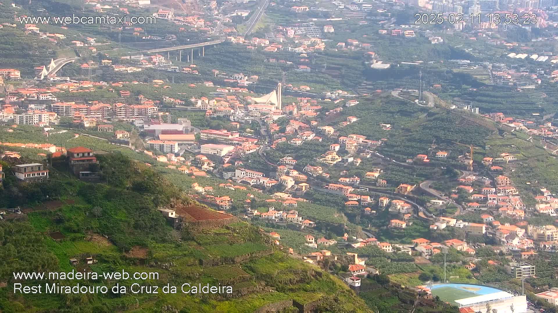 A hazy, high-angle view shows a hillside town with numerous orange-roofed houses nestled among green vegetation and winding roads, including a large, visible bridge in the distance.