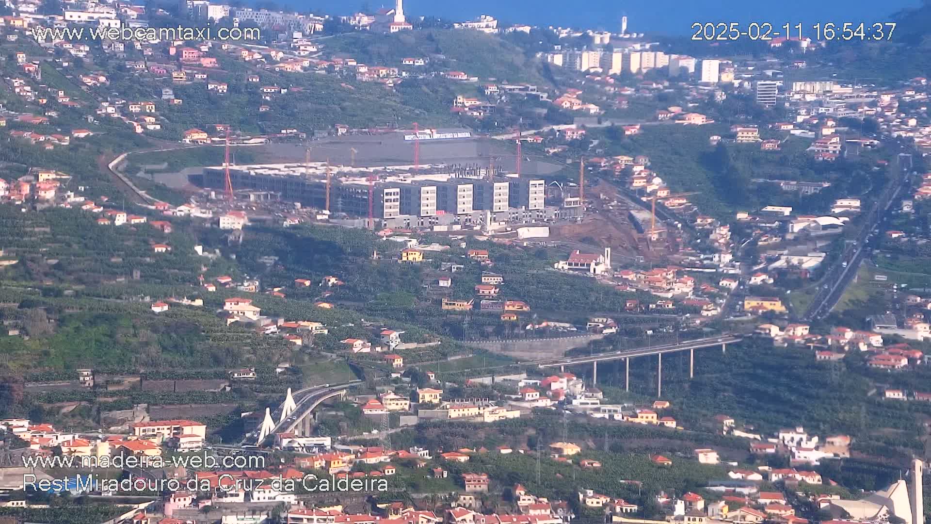 A high-angle, long shot shows a coastal town on a hillside under a clear sky, with a large building under construction and a bridge visible in the foreground.