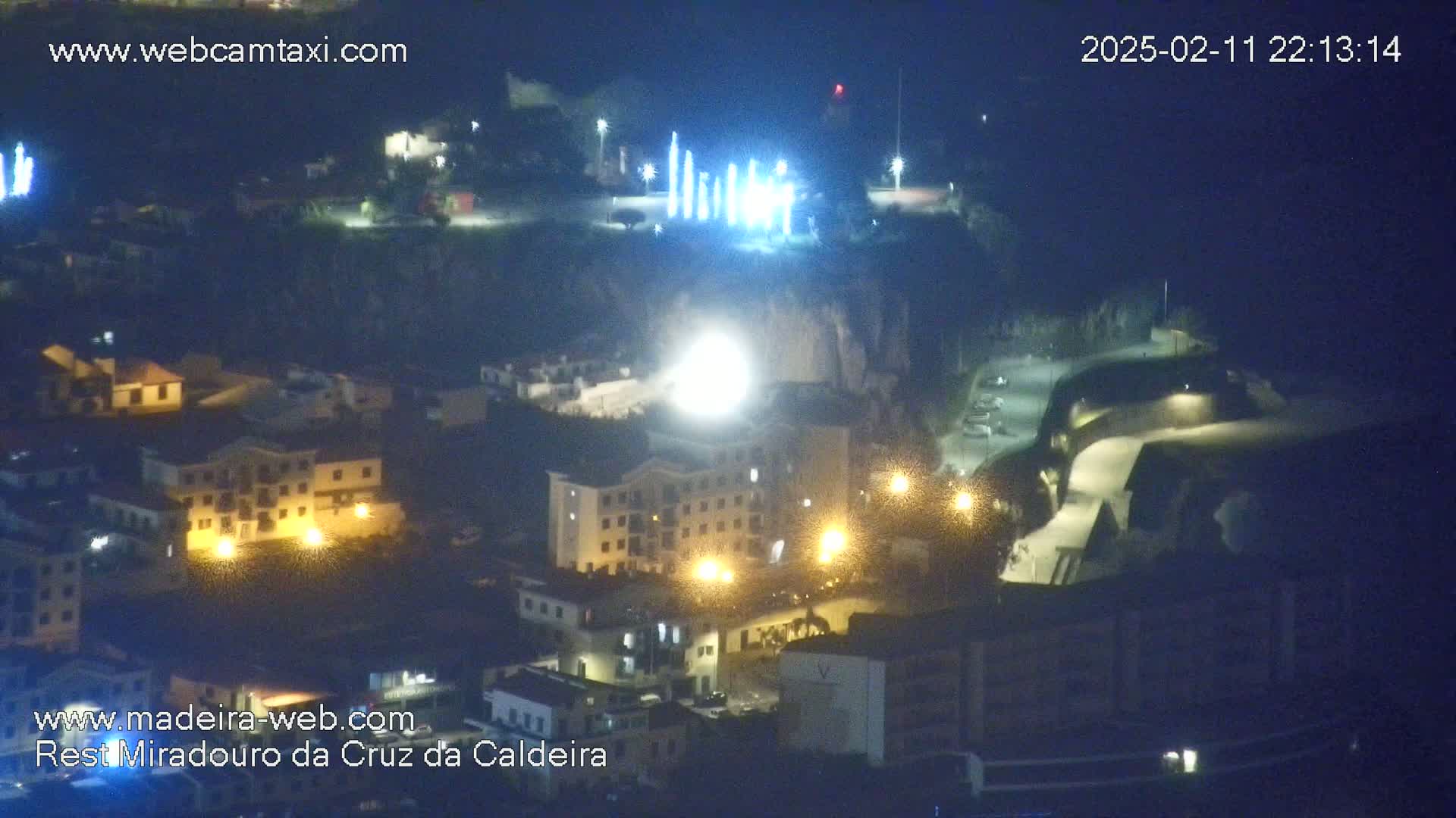 A nighttime aerial view of a town illuminated by streetlights, with a brightly lit area in the center and what appears to be a light display on a hill in the background.