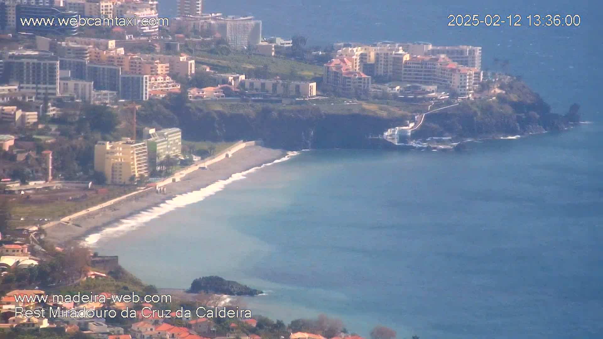 A dark-sand beach with gentle waves laps against a rocky coastline near a cluster of buildings under a clear sky.
