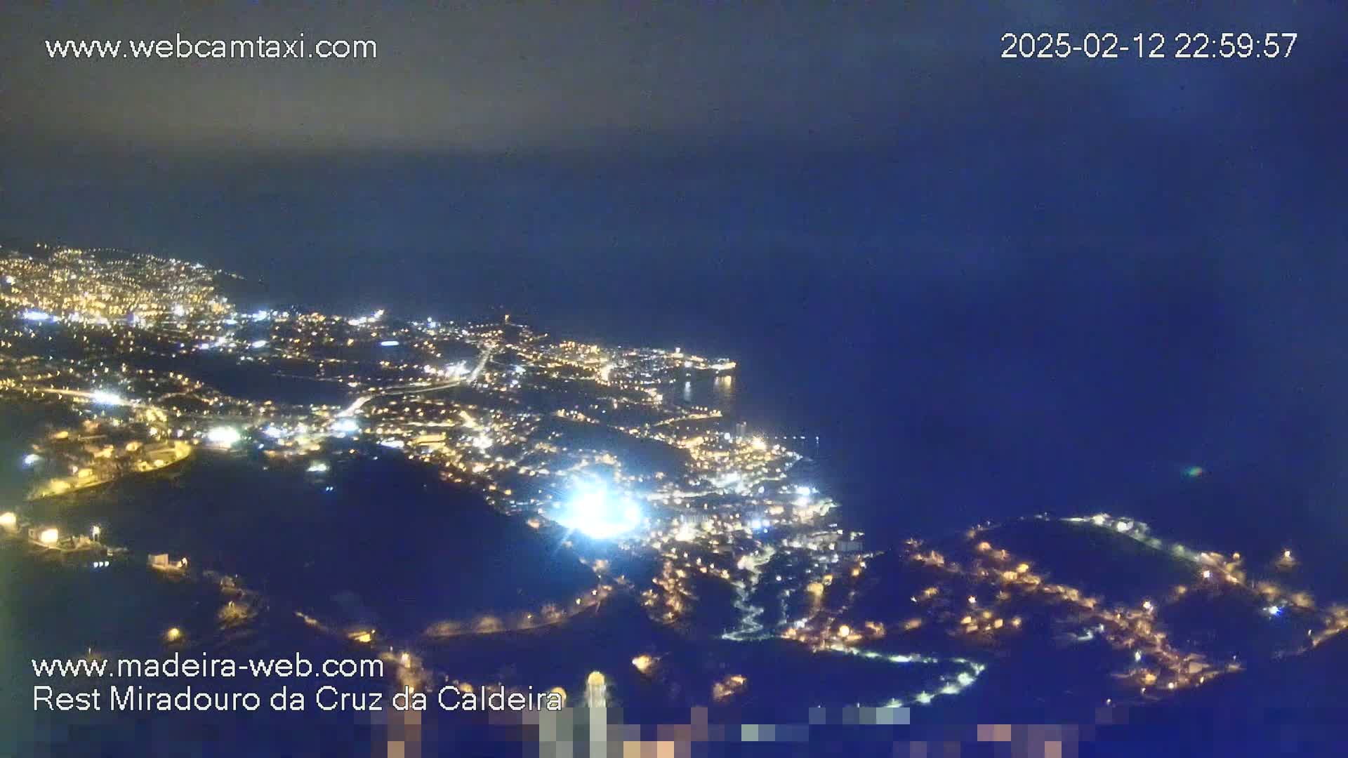 A nighttime aerial view shows a coastal city illuminated by many lights, under a dark, clear sky.