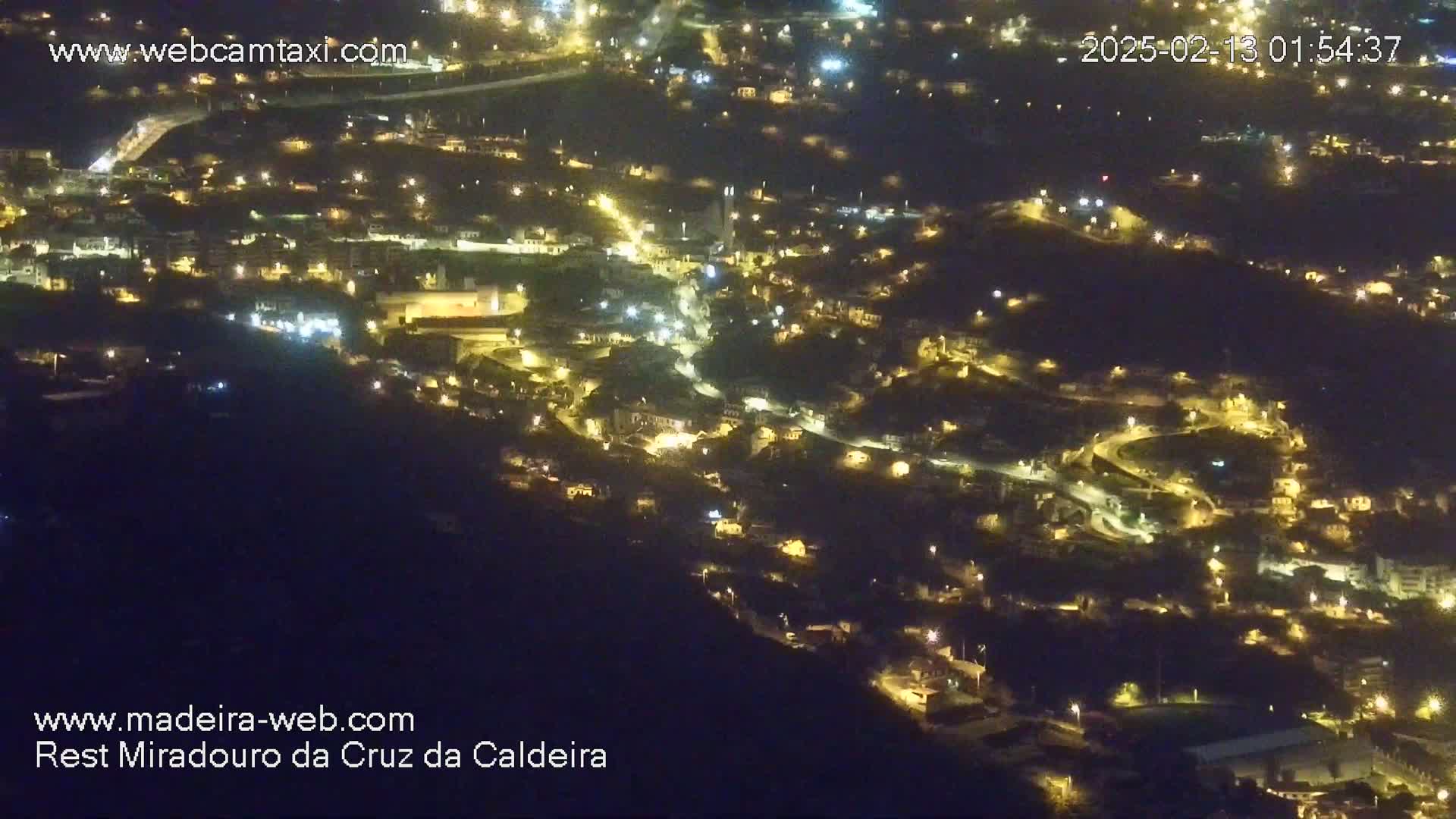 A nighttime, aerial view of a town nestled on a hillside, illuminated by numerous lights.