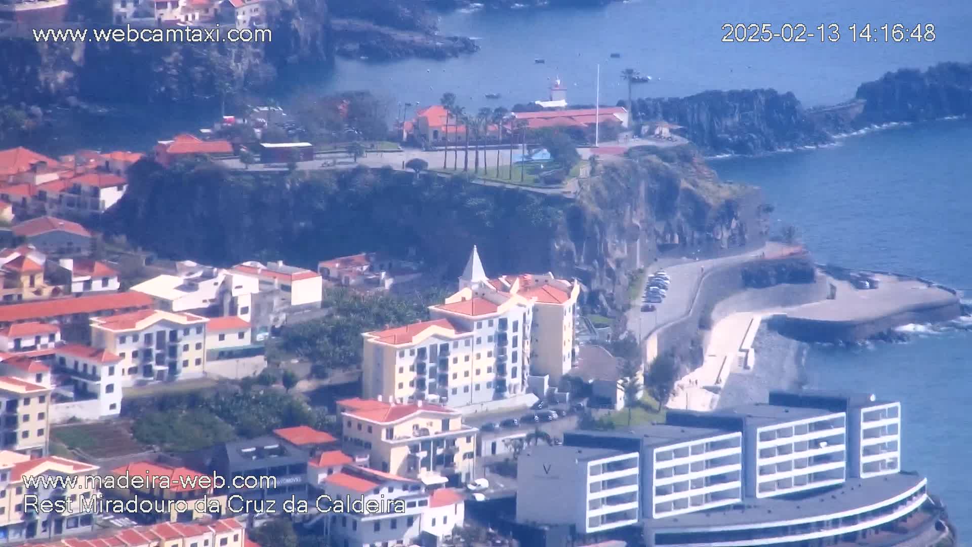 An aerial view shows a coastal town built on a cliffside overlooking a calm ocean on a sunny day.