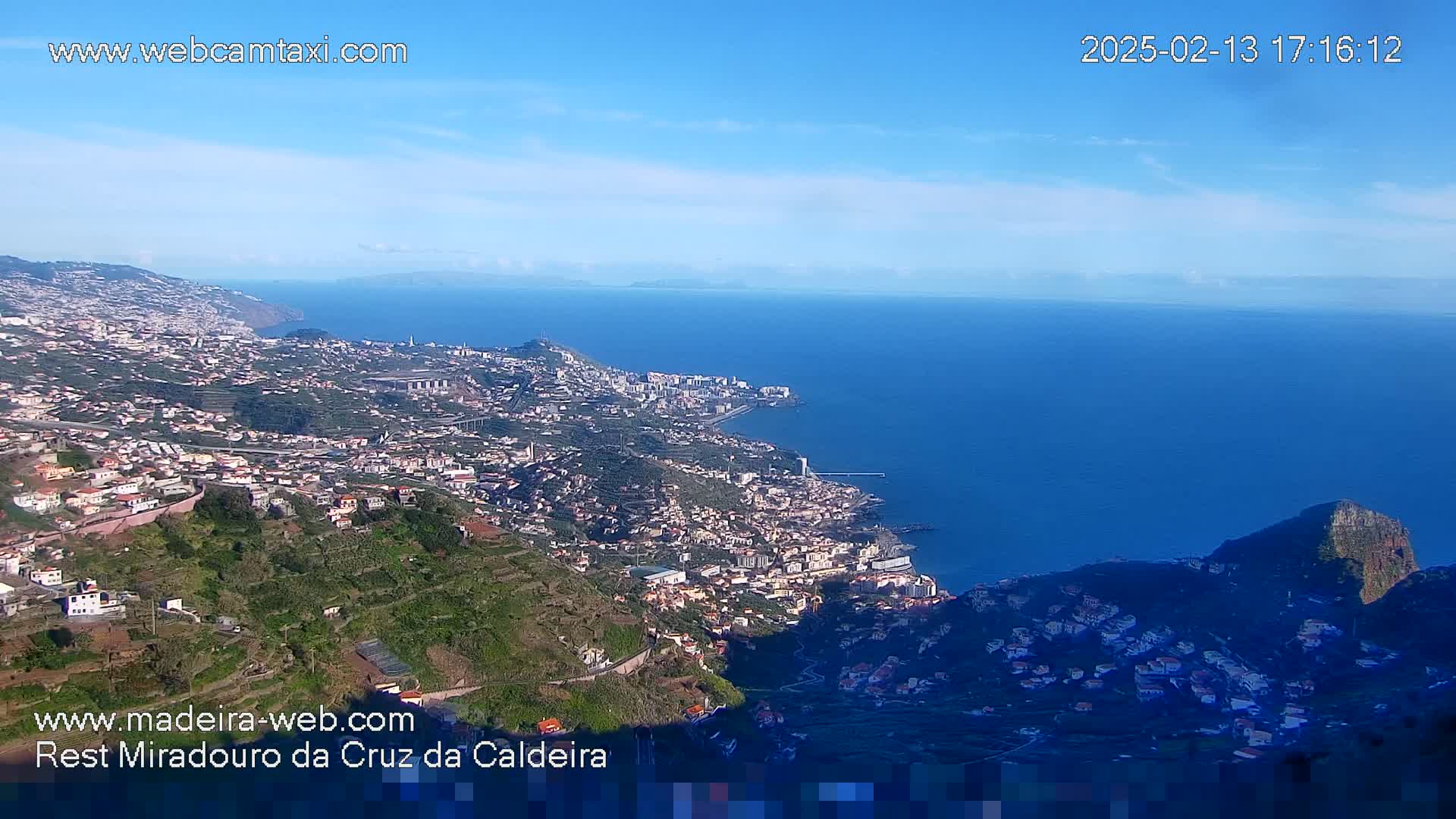 A coastal city nestled in a mountainous region overlooks a vast blue ocean under a clear, bright sky.