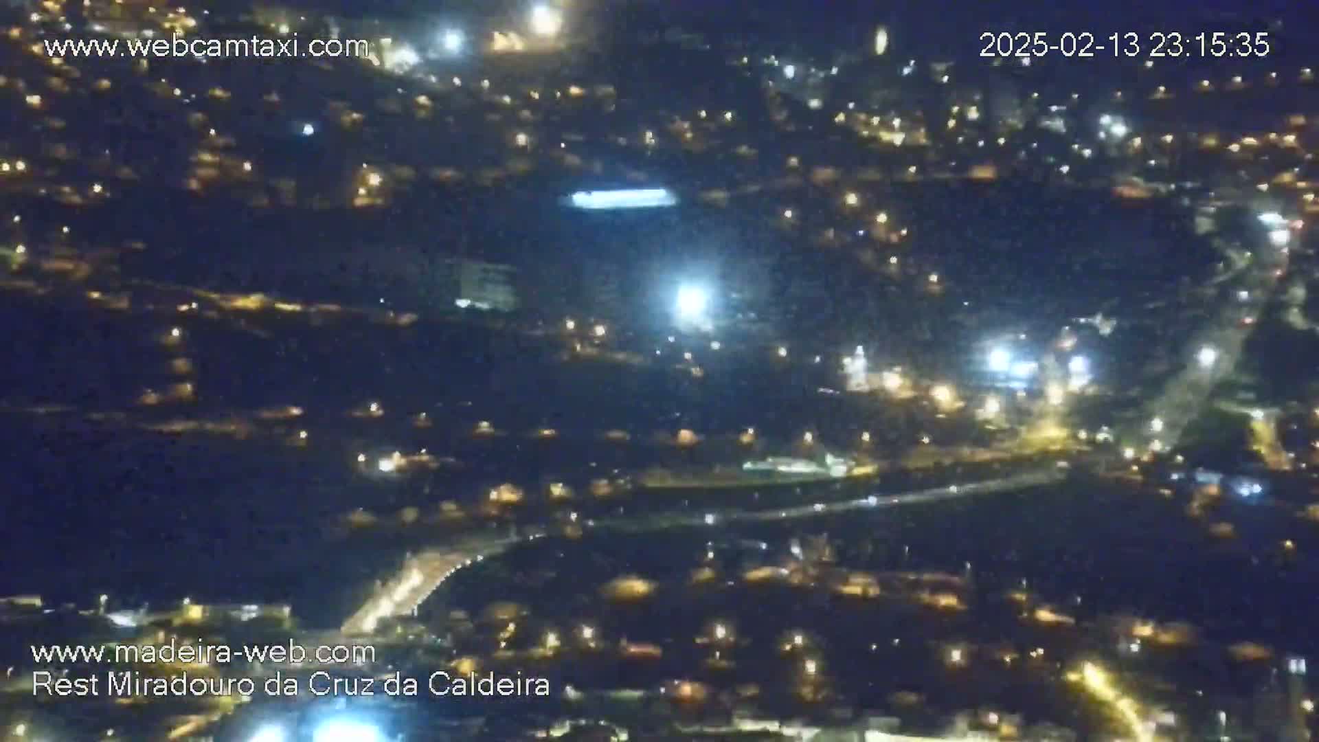 A nighttime aerial view shows a sprawling town illuminated by numerous lights, with a highway curving through the landscape.