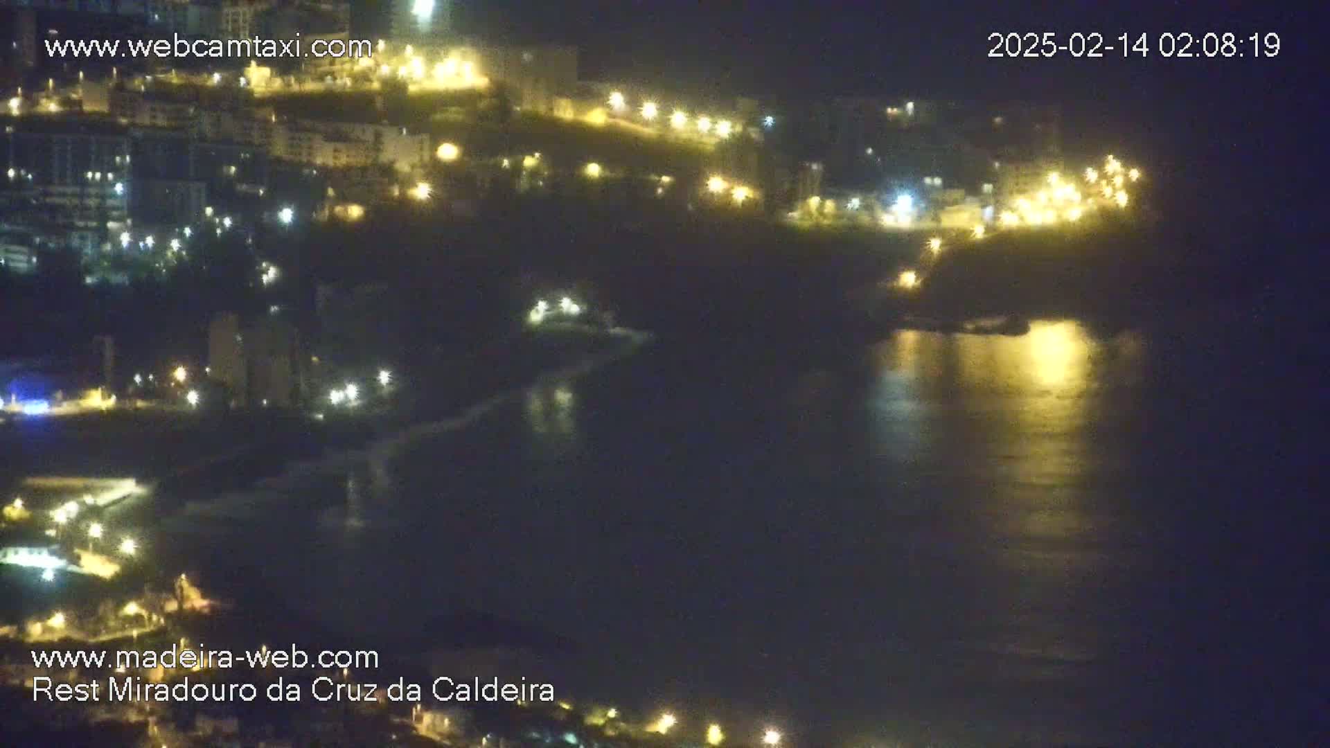 A nighttime coastal scene shows a dark body of water reflecting the lights of a town built on a hillside.