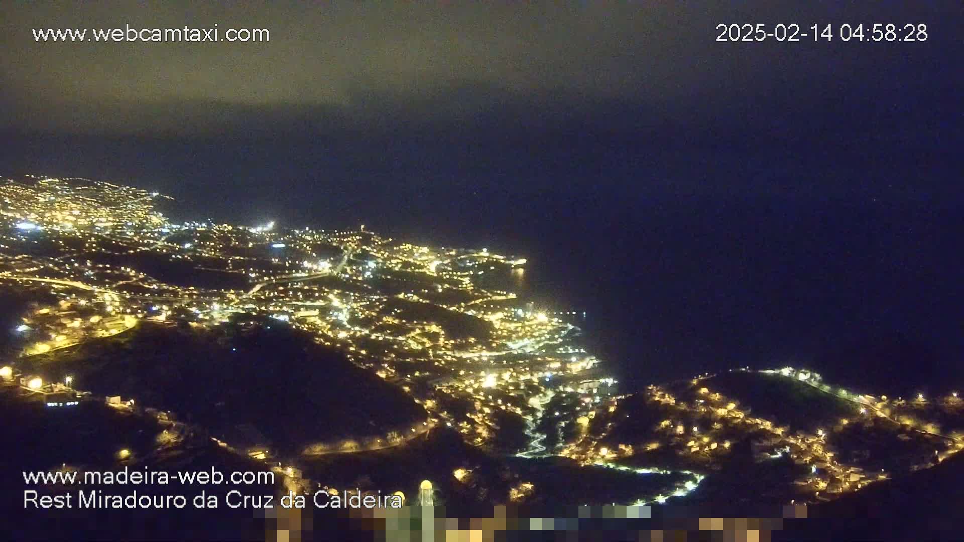 A nighttime, aerial view shows a coastal city illuminated with many lights, stretching along a dark ocean under a cloudy sky.