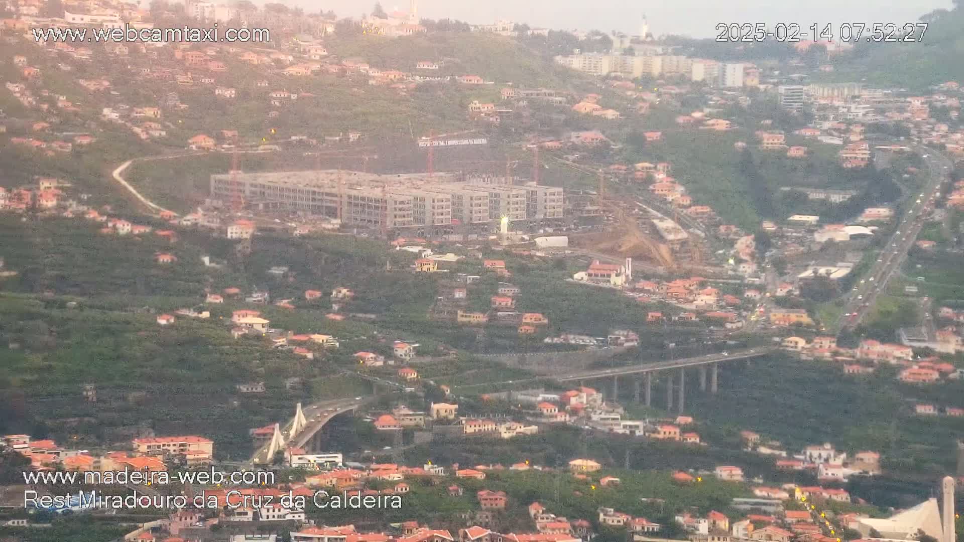 A hazy, high-angle view shows a large building under construction nestled among many smaller buildings on a hillside, with highways and bridges weaving through the landscape.