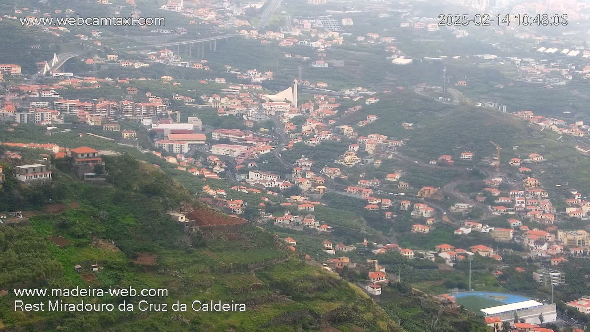A hazy, high-angle view shows a hillside town with many red-roofed buildings, terraced farmland, and a large bridge in the distance.