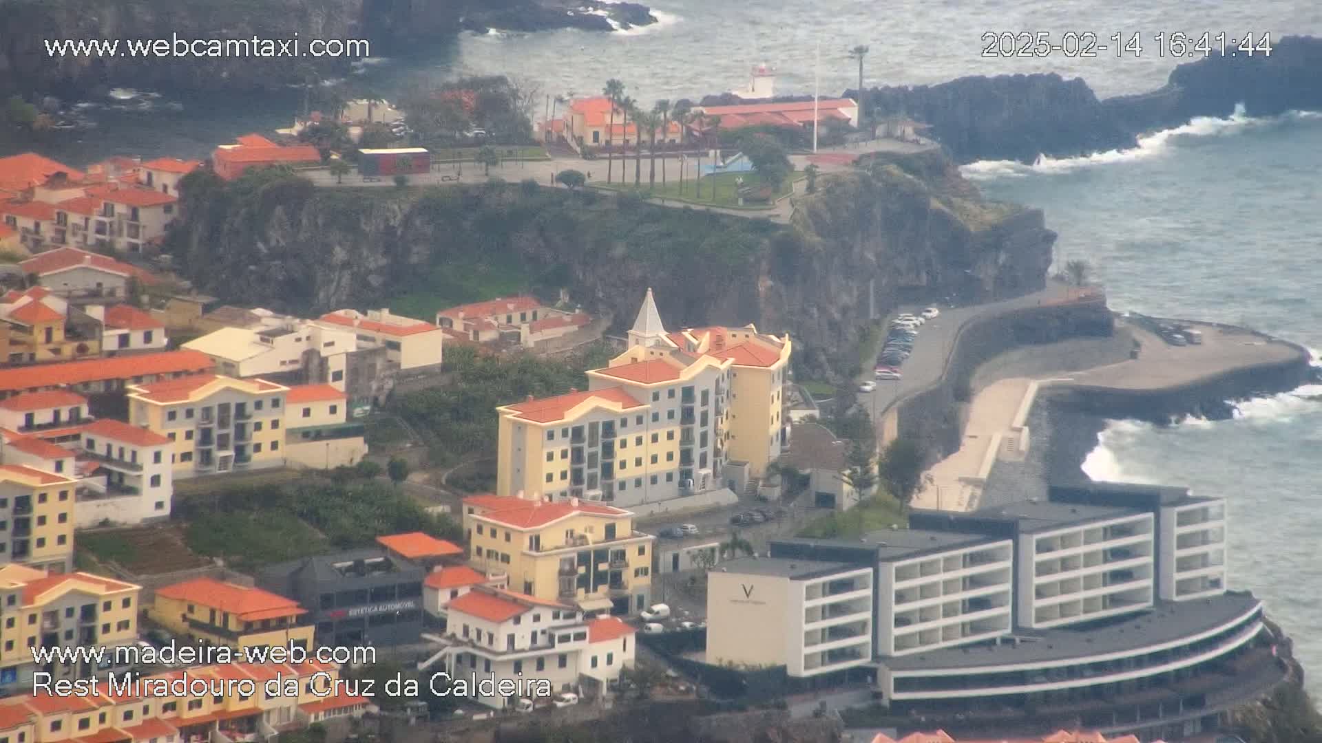 An aerial view shows a coastal town nestled against a cliffside, with buildings of varying heights and colors, and a dark, rocky shoreline with waves crashing against it.