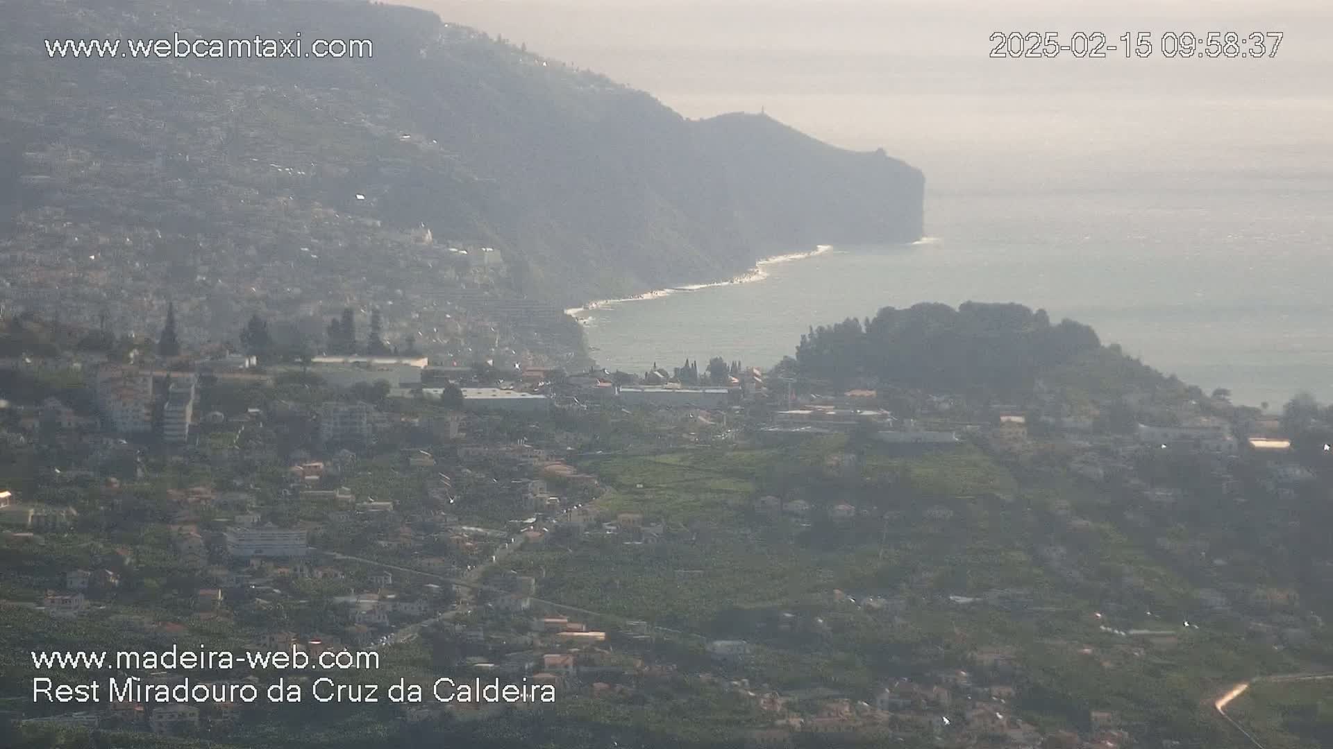 A hazy coastal town nestled between a steep cliff and calm ocean is seen from a high vantage point.