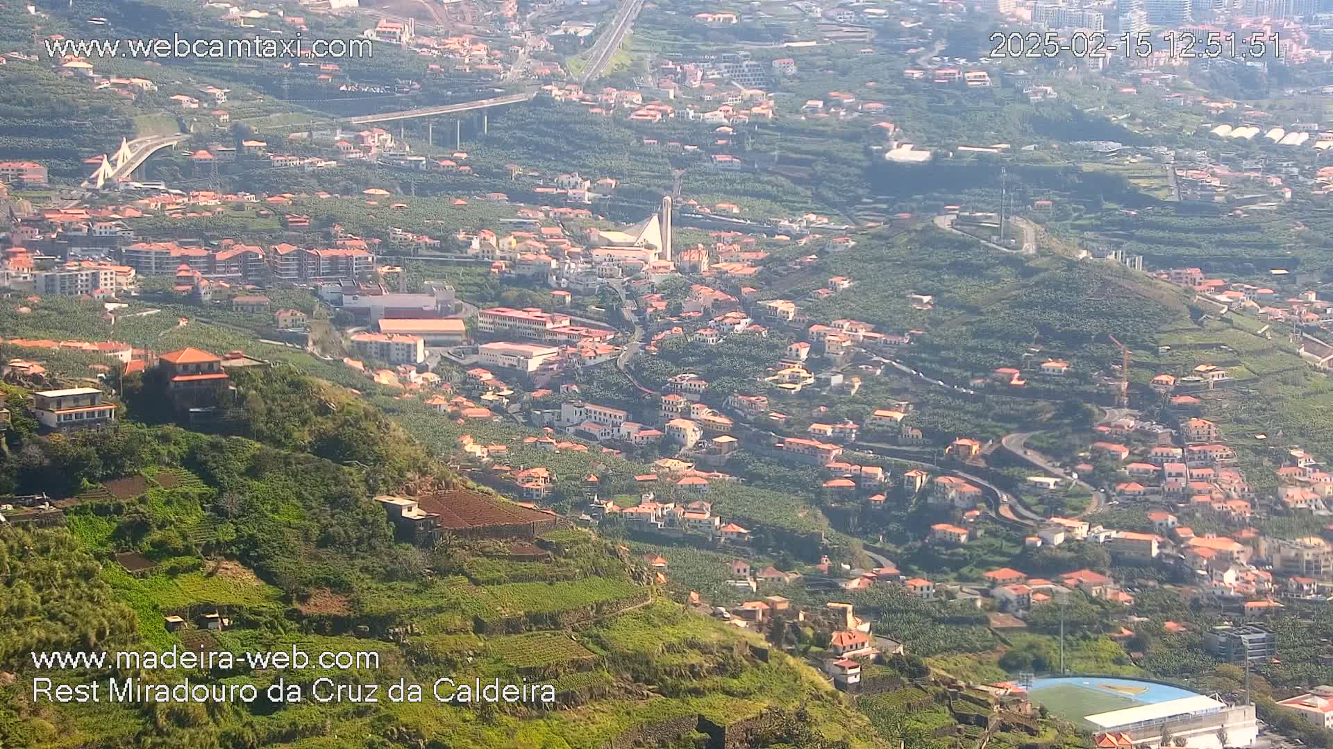 A hillside town with red-roofed houses and terraced farmland, seen under sunny conditions, stretches down to a small soccer field.