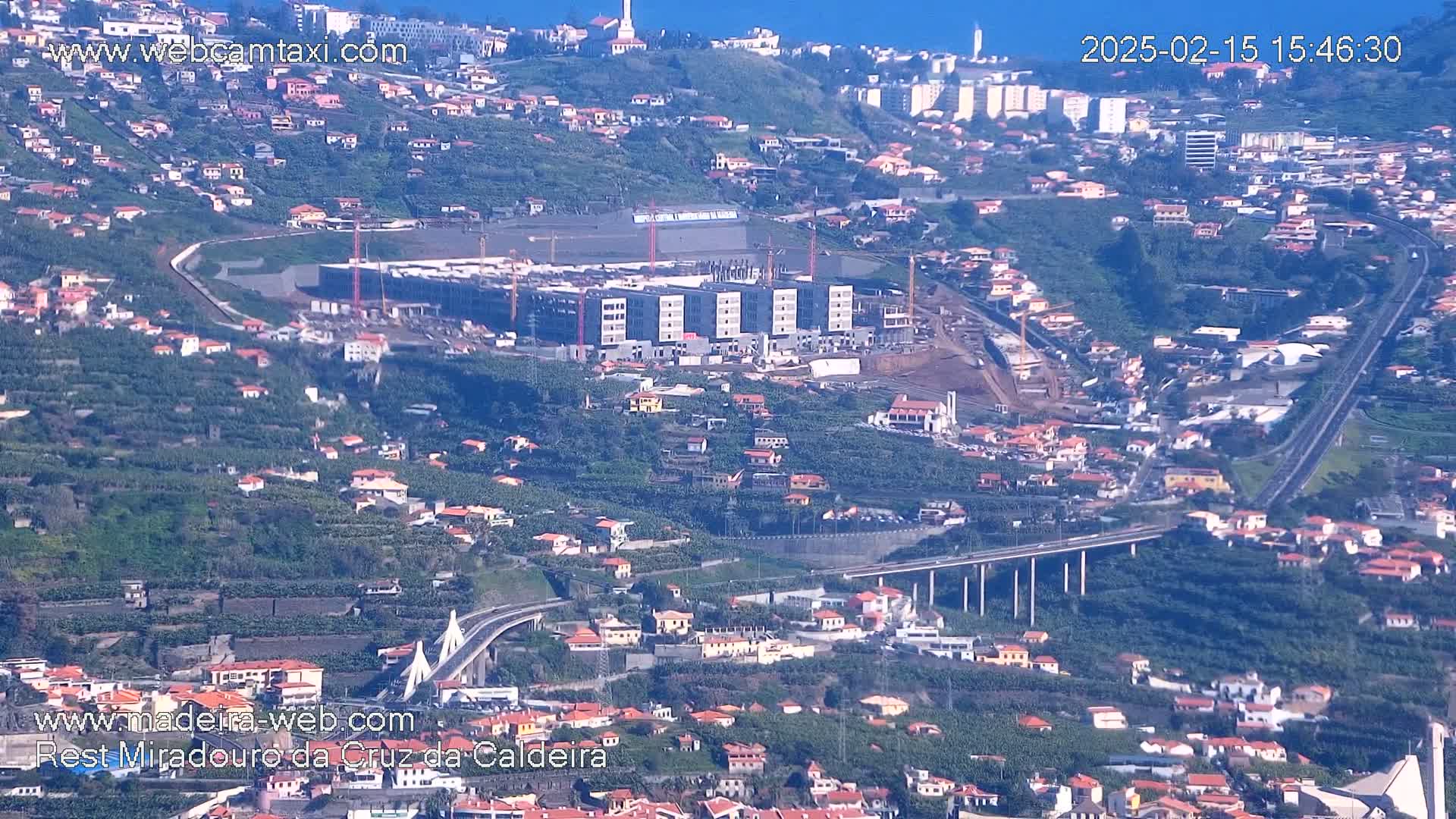 An aerial view shows a hillside town with a large building under construction and a bridge spanning a roadway.