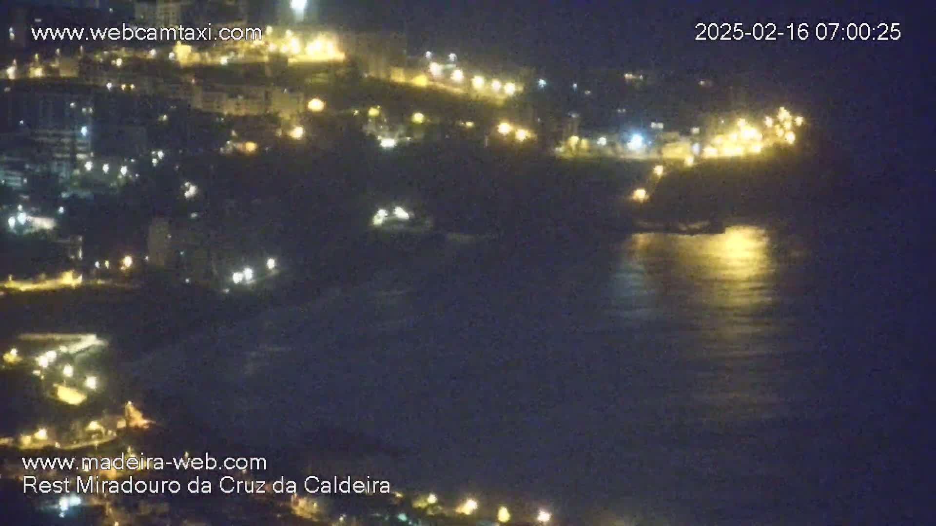 A nighttime coastal scene shows a dark body of water reflecting lights from a hillside town.