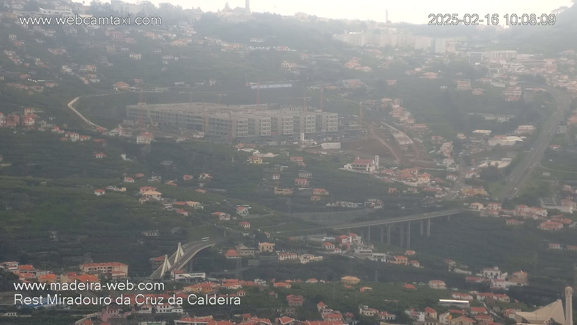 A hazy view of a hillside town with a large building under construction, a bridge, and a highway.