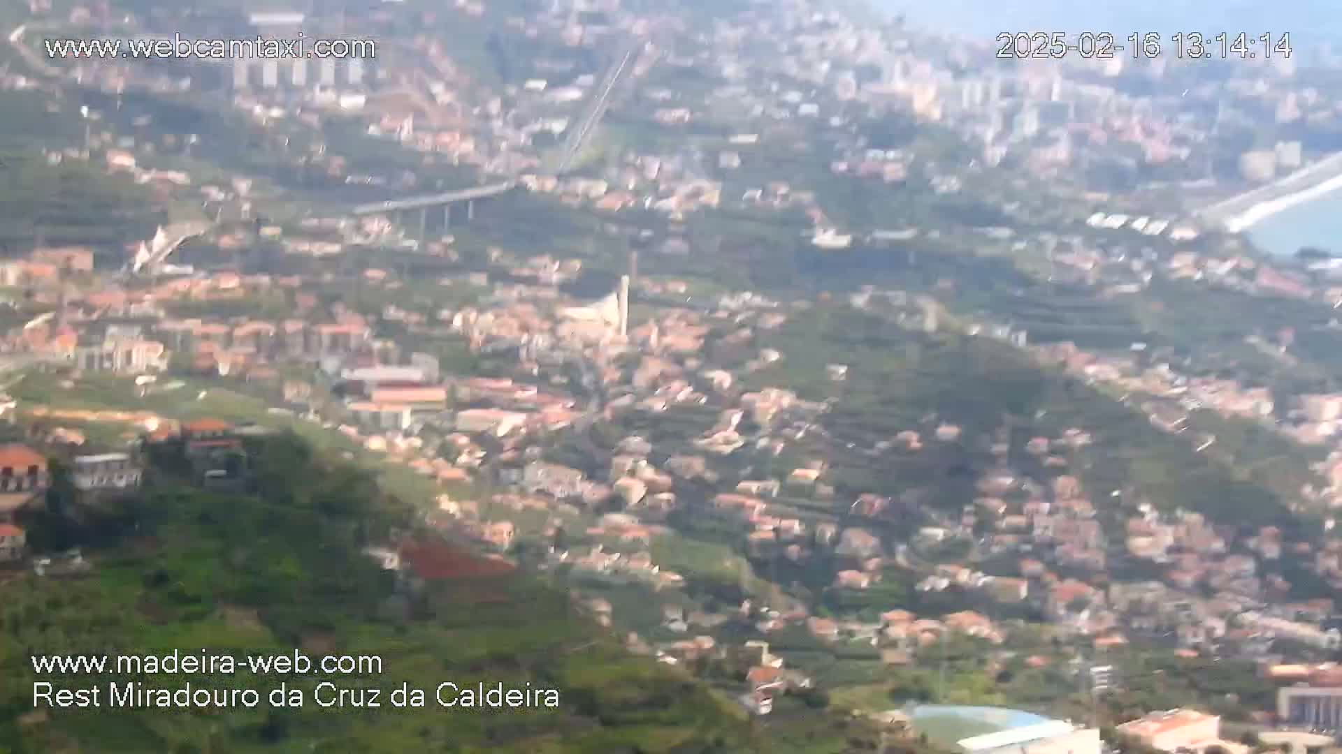 A hazy, high-angle view shows a sprawling town nestled in a verdant hillside, with a bridge and a body of water visible in the distance.