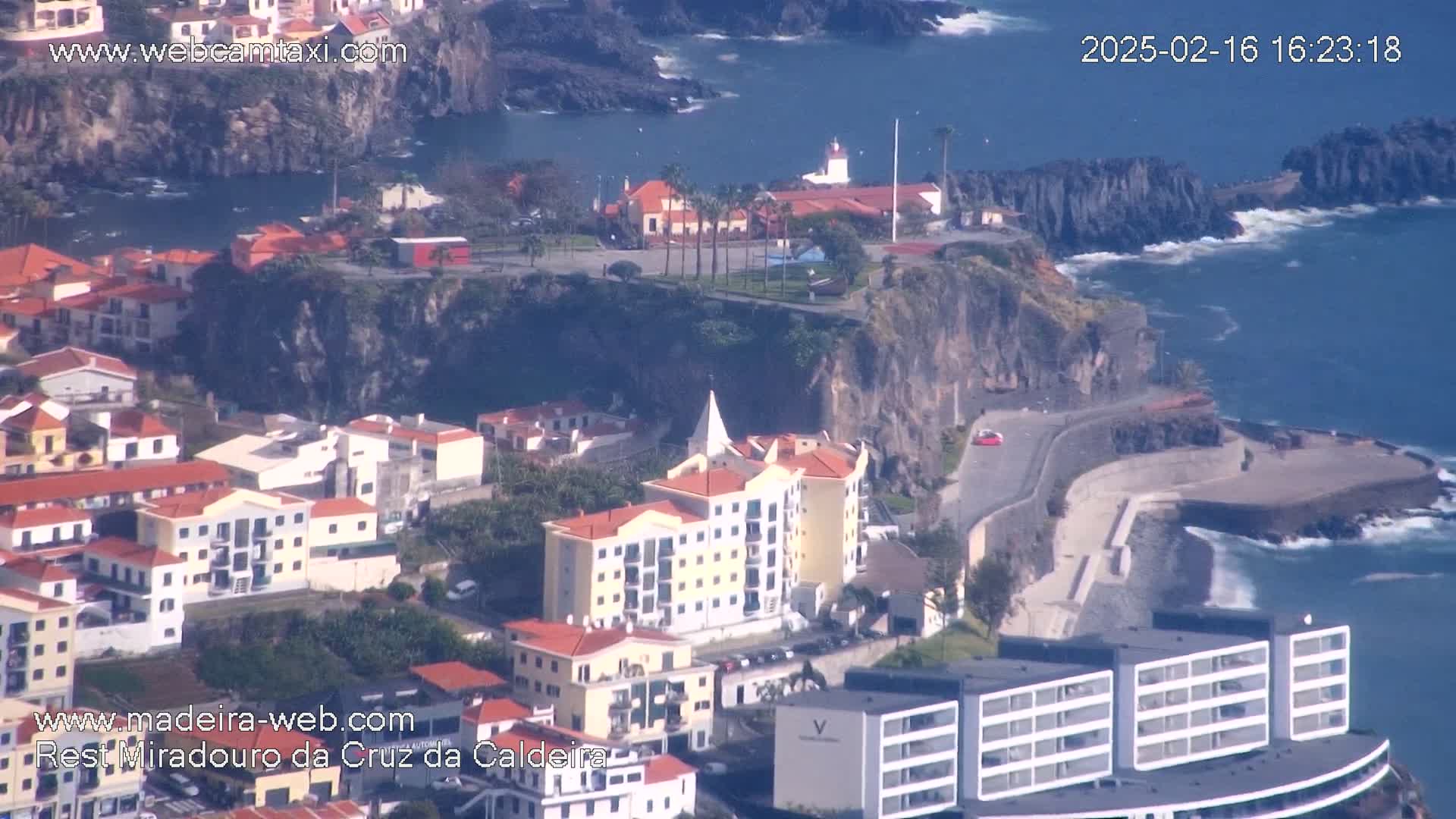 An aerial view shows a coastal town nestled against dark cliffs, with a calm ocean and some small waves crashing against the rocks.