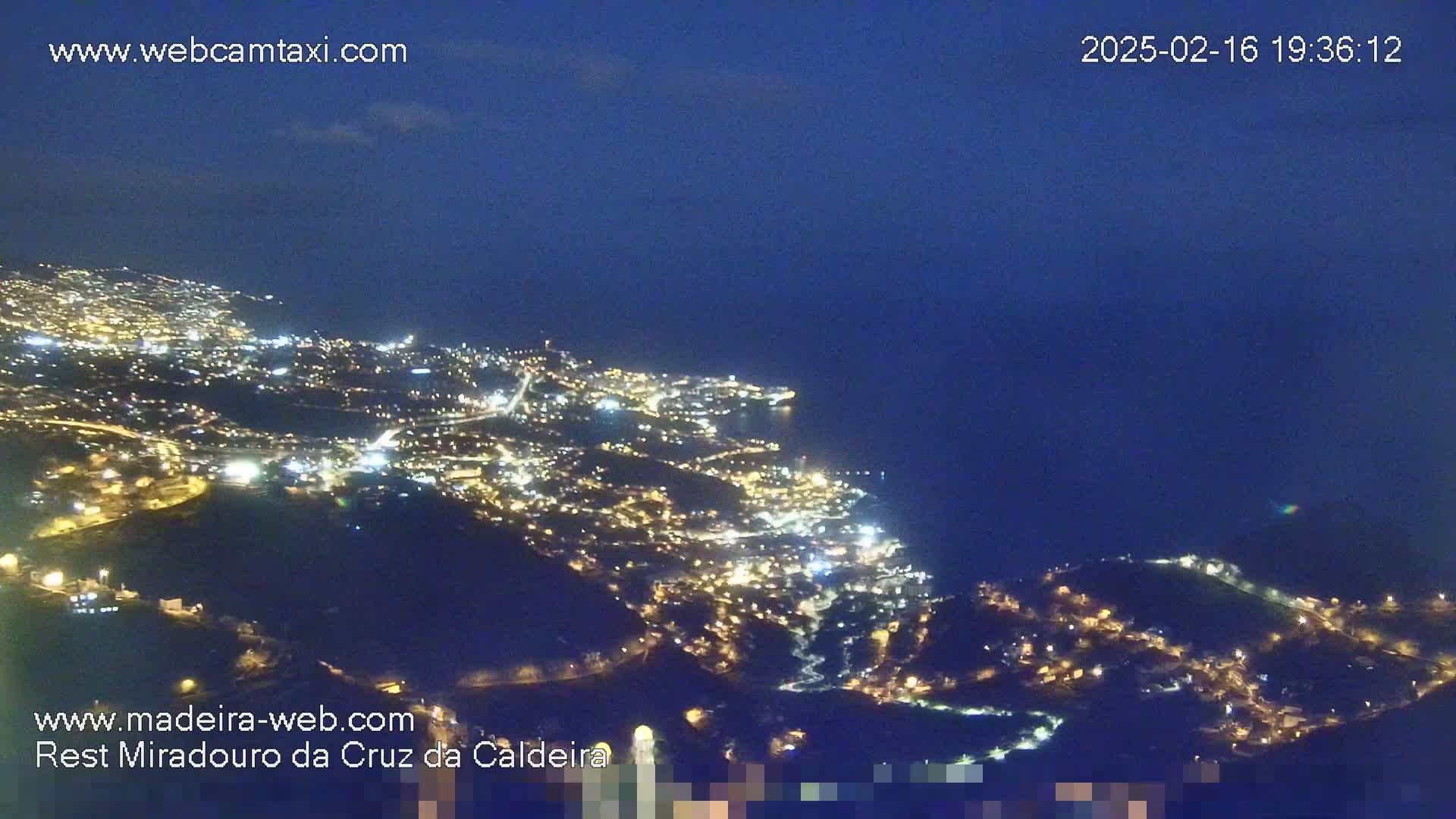 A nighttime aerial view shows a coastal city illuminated with lights, stretching along a dark ocean under a clear, dark blue sky.