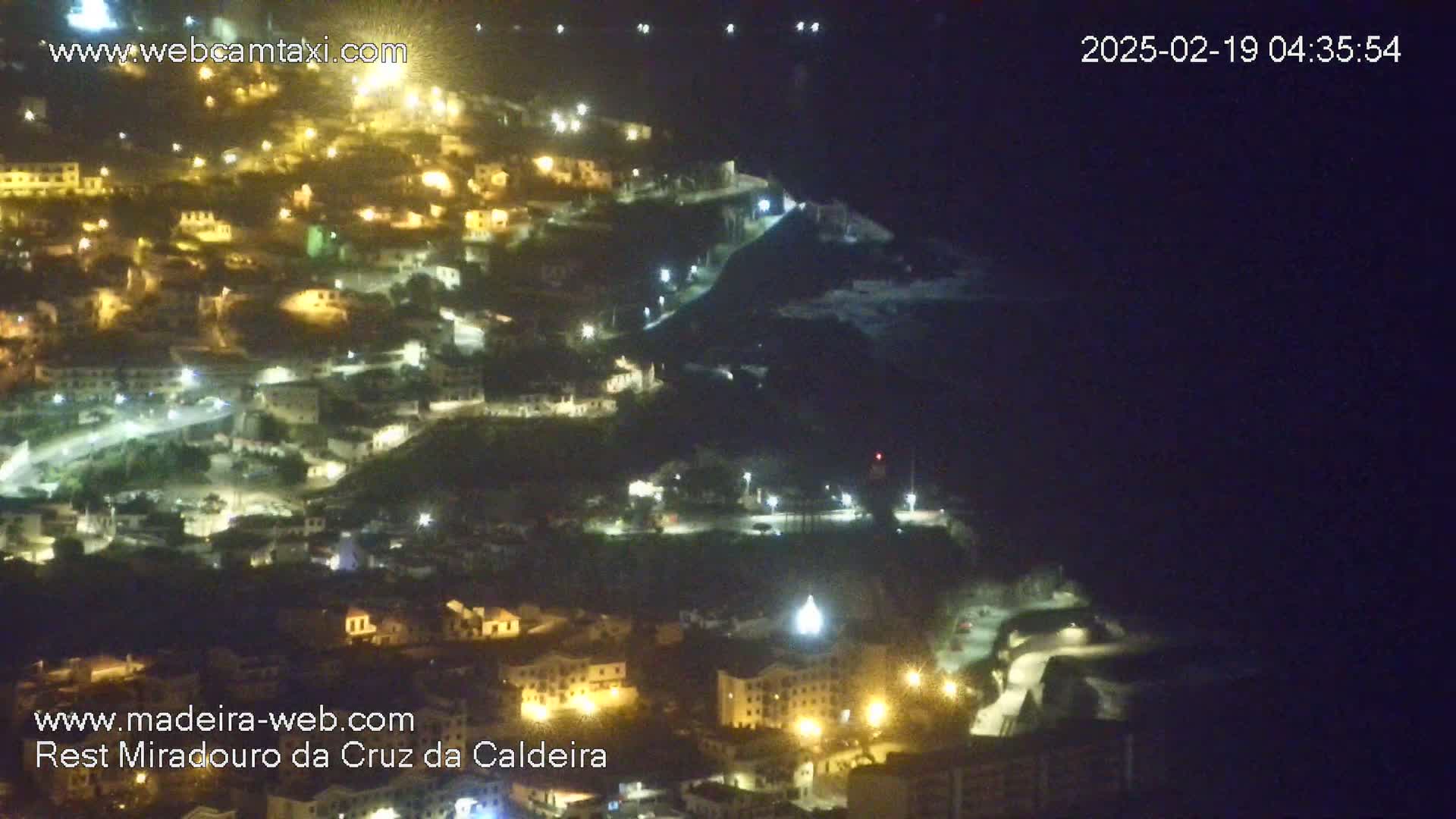 A nighttime, aerial view shows a hillside town illuminated by numerous lights, extending down toward a dark body of water.