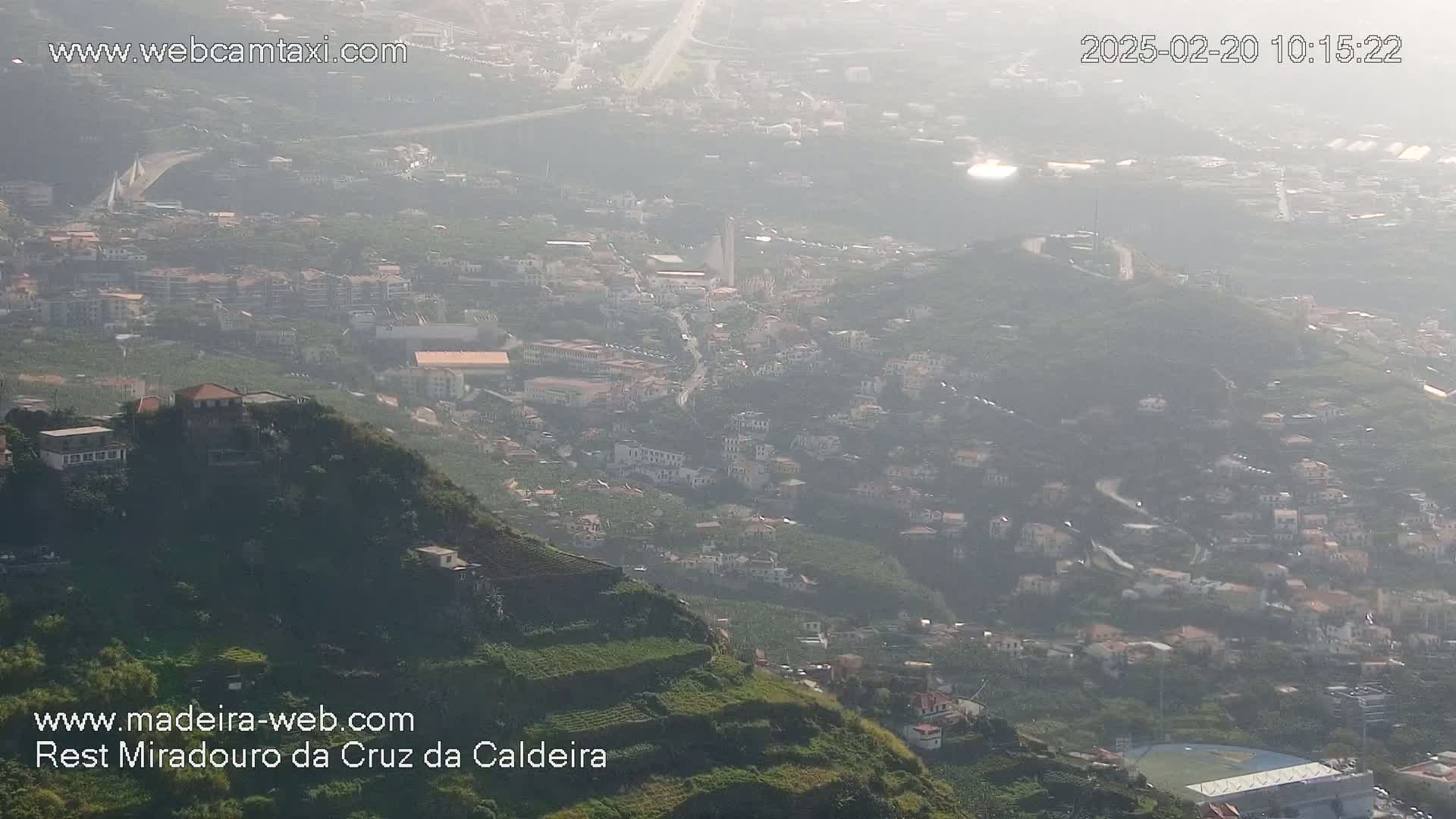 A hazy, high-angle view shows a hillside town with numerous houses, terraced farmland, and a distant bridge under a light haze.