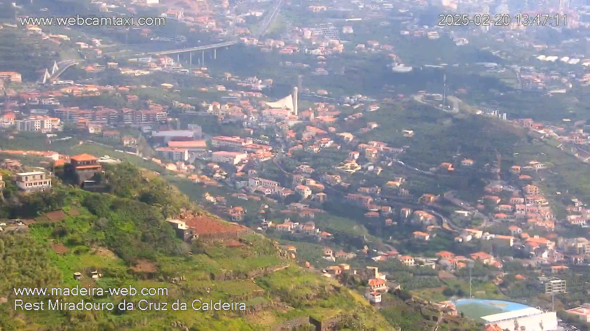 An aerial view shows a hillside town with many low-rise buildings under hazy, sunny conditions,  a bridge visible in the distance.