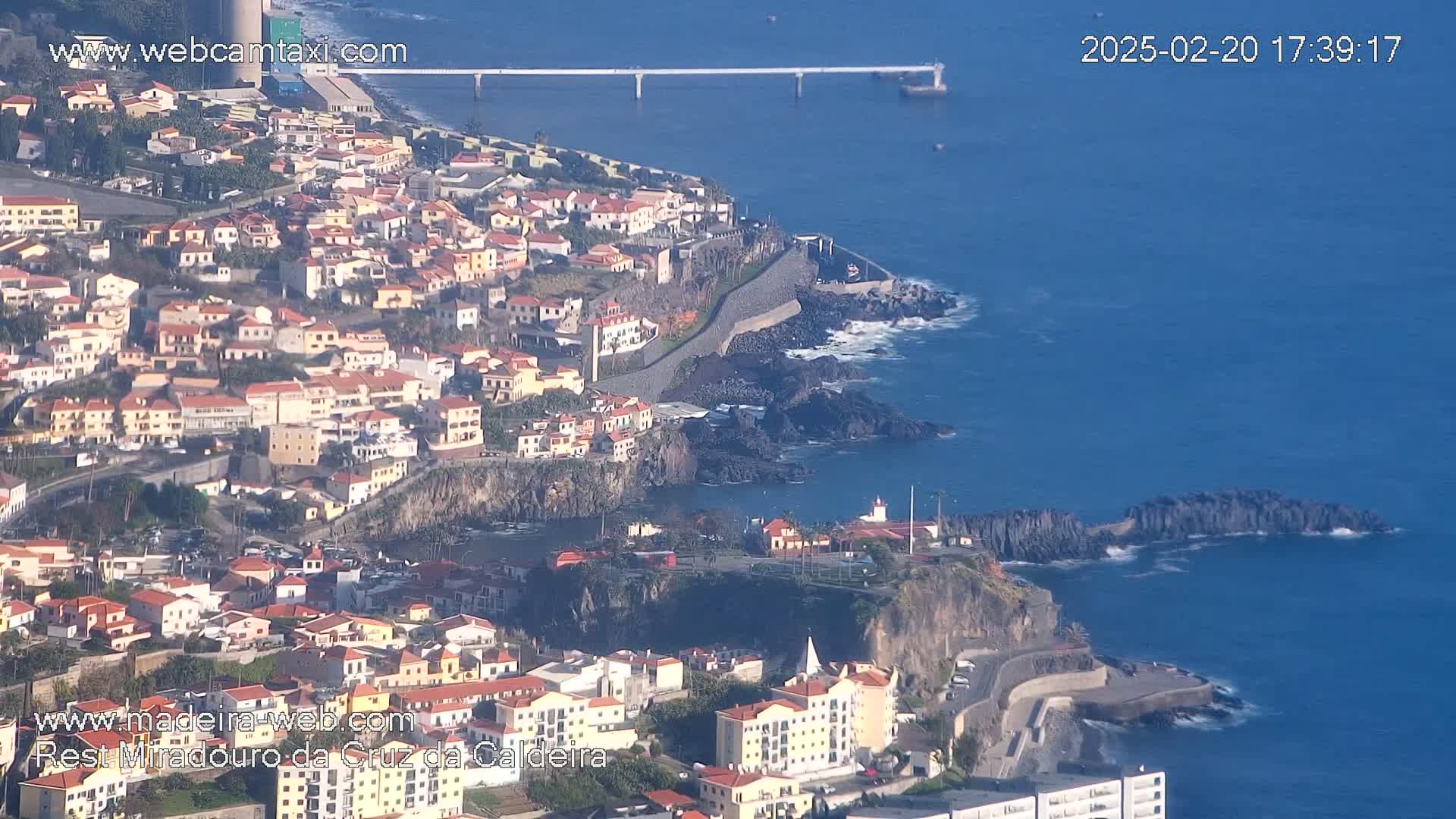 A coastal town with numerous low-rise buildings clings to a rocky cliffside overlooking a calm, deep blue ocean on a sunny day.