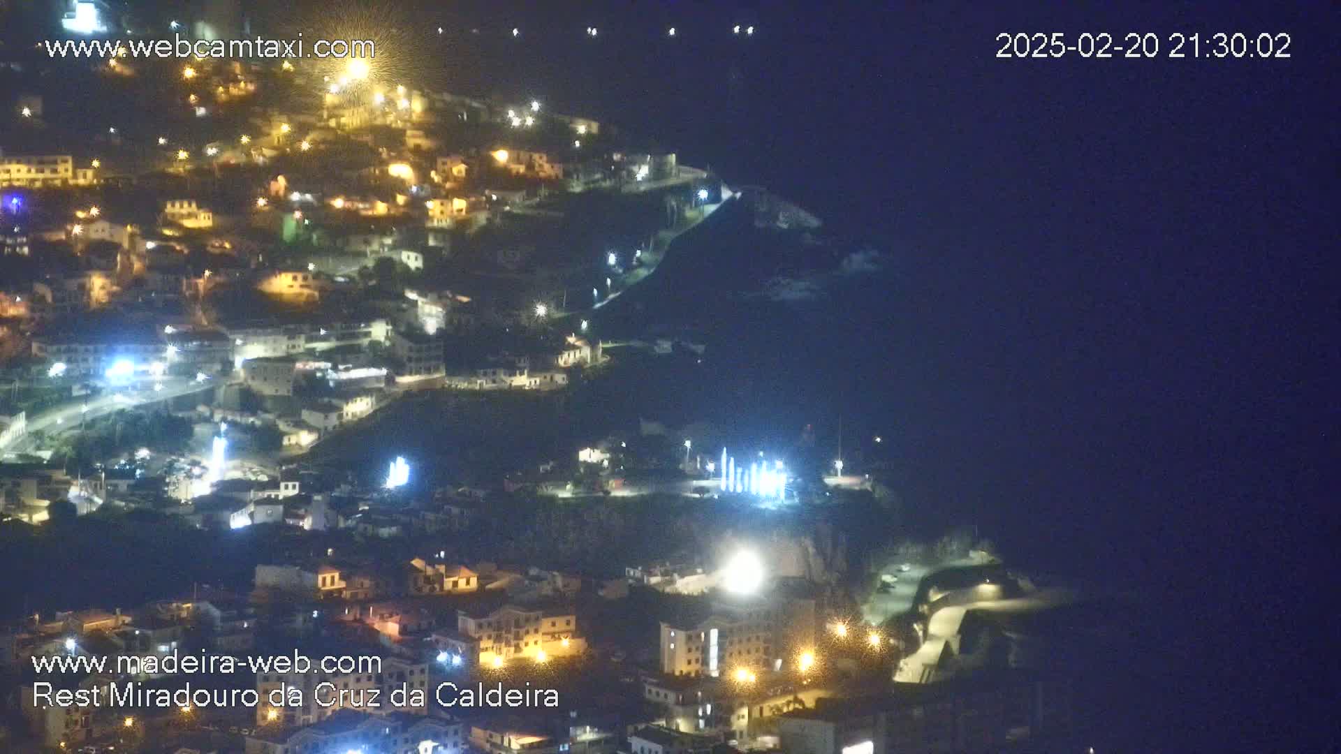 A nighttime, aerial view of a brightly lit town nestled on a hillside overlooking a dark body of water.