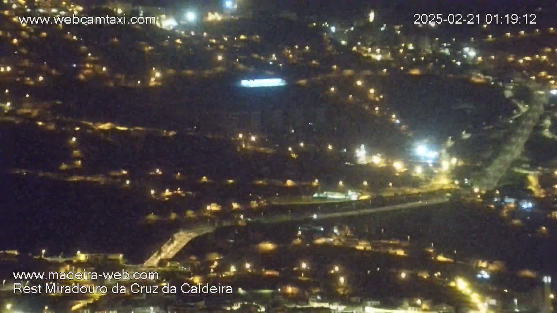 A nighttime aerial view shows a town illuminated by numerous lights spread across a hilly landscape.