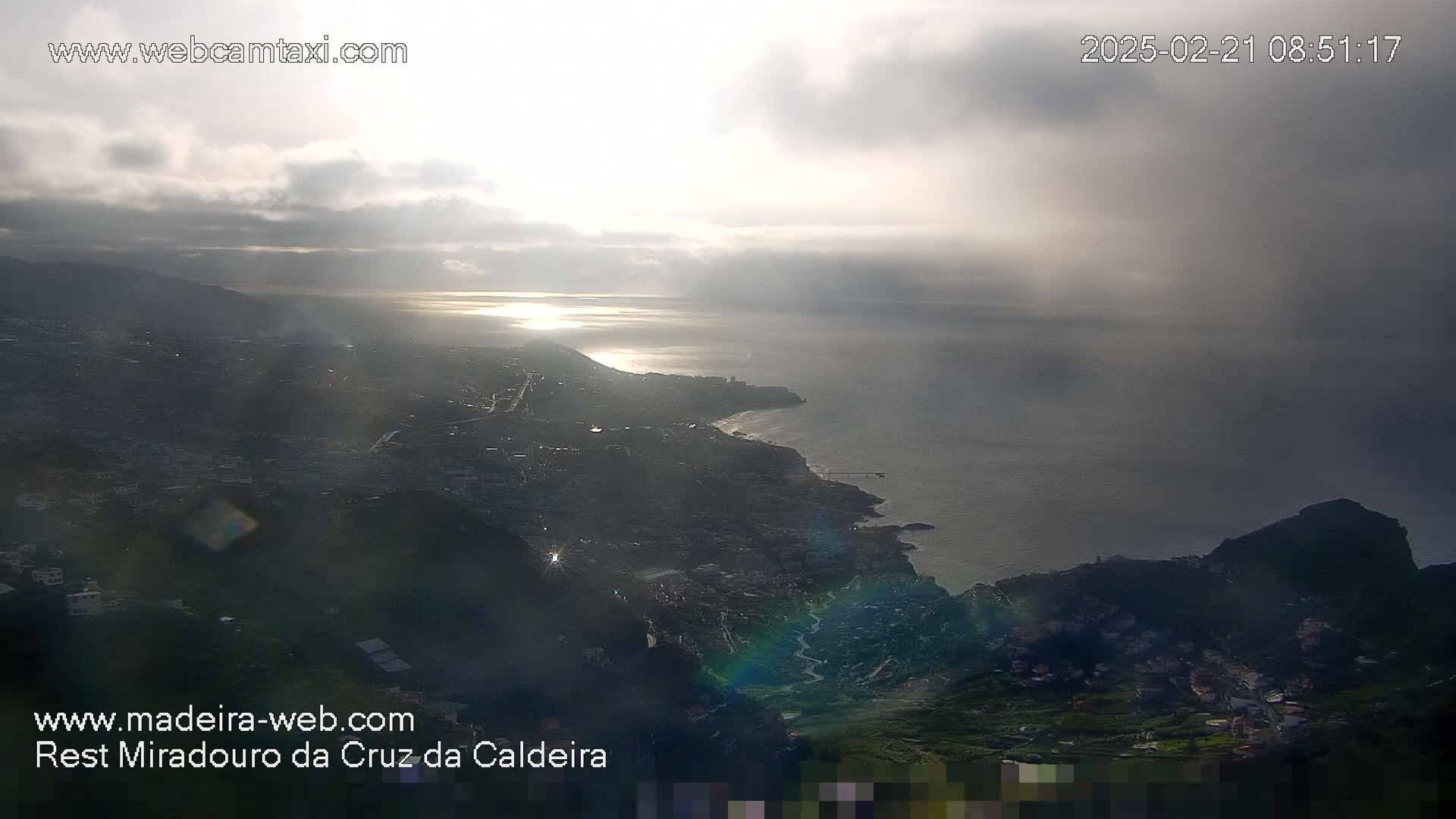 A coastal city nestled between mountains and the ocean is seen under a cloudy sky with sunlight breaking through.