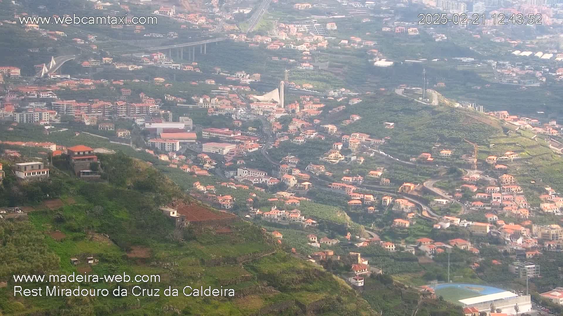 A hazy view shows a hillside town with red-roofed buildings, a large bridge in the distance, and a sports field below.