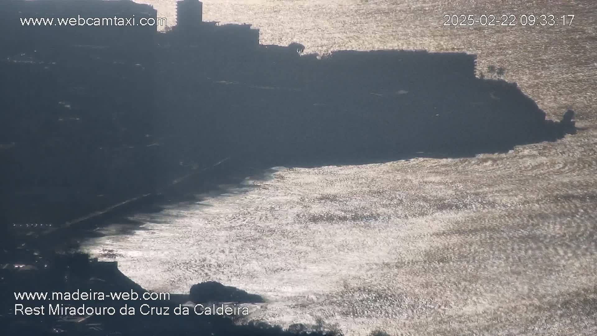 A silhouetted coastal cliff and buildings overlook a bright, sparkling ocean under a mostly sunny sky.