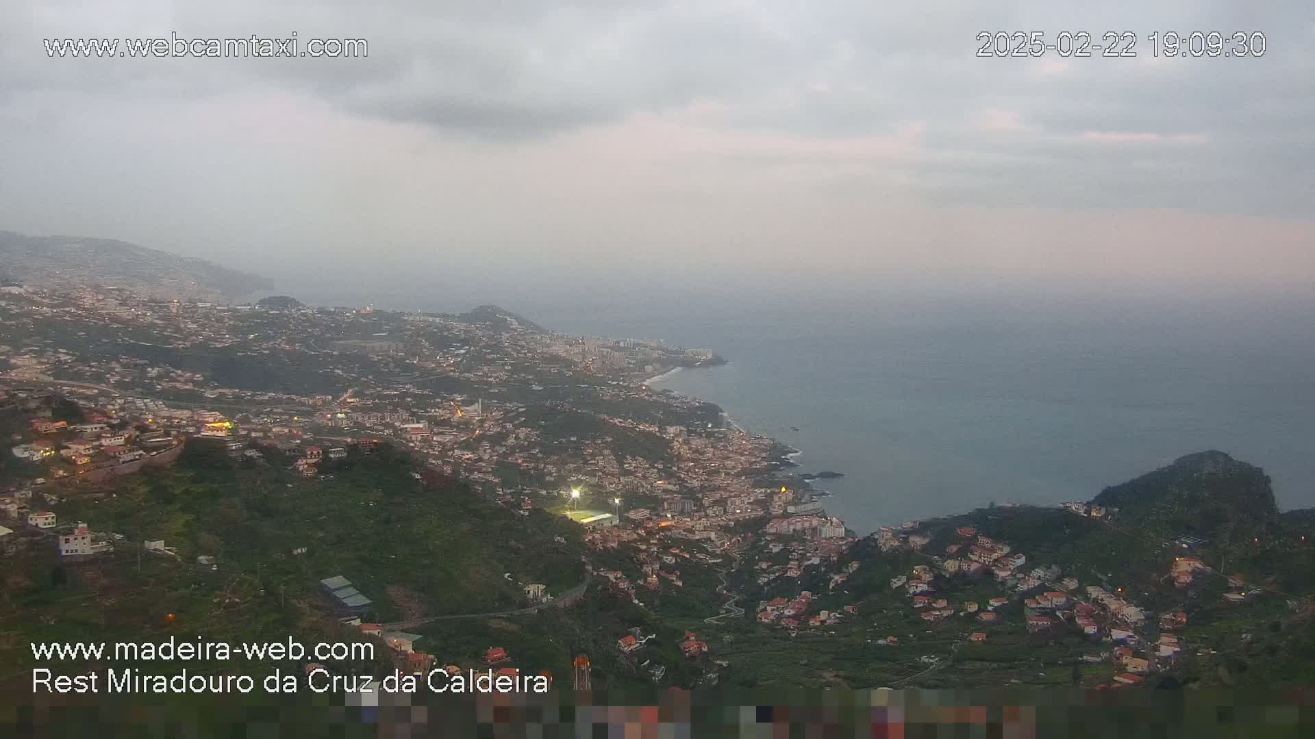 A coastal town at dusk under a cloudy sky is viewed from a high vantage point, showing the town's lights illuminated against the dark green hills and ocean.