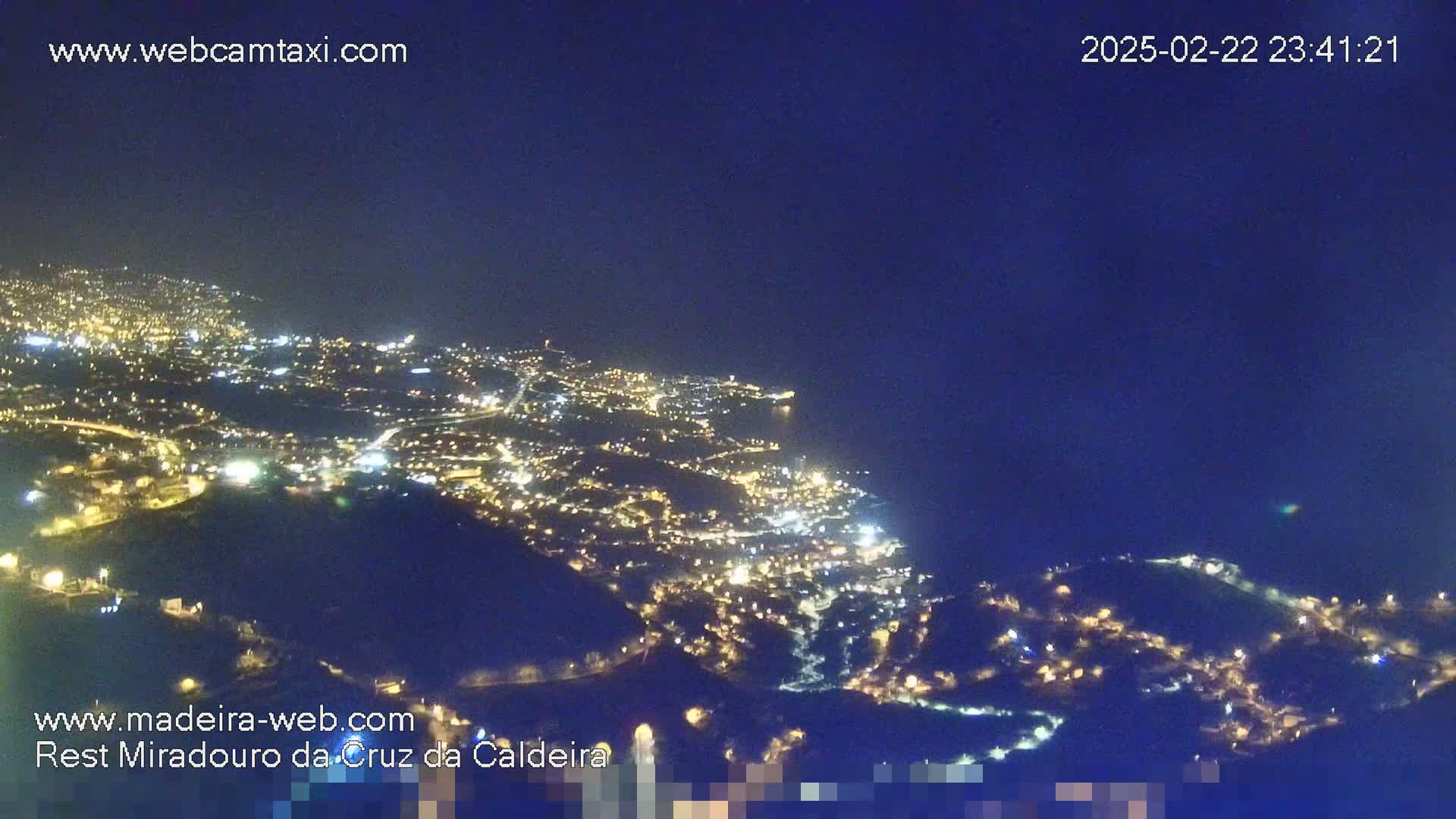 A nighttime, high-angle view of a coastal city illuminated by numerous lights against a dark blue sky.