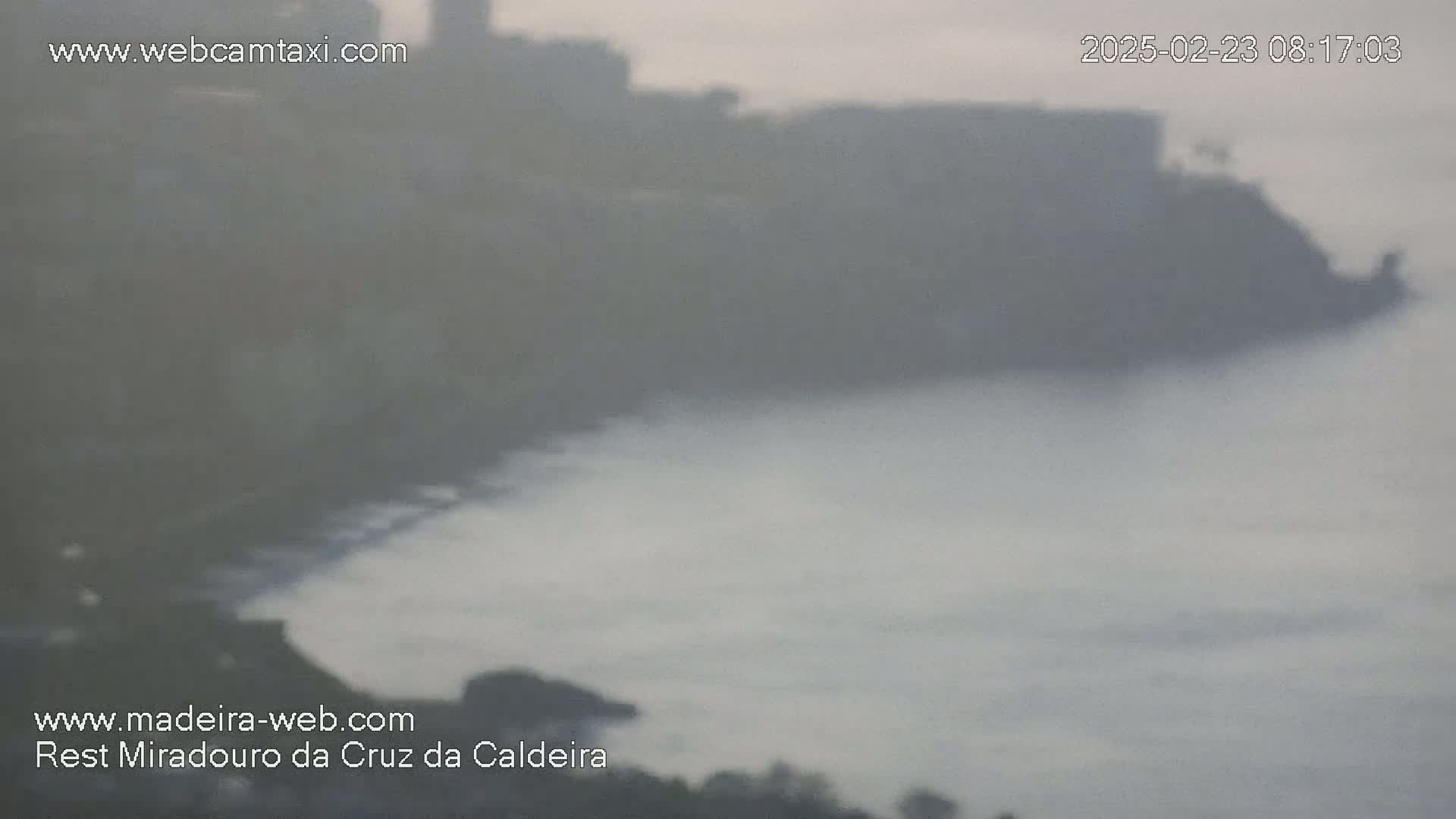 A hazy, overcast day reveals a coastline with buildings and a dark-colored beach meeting a calm, gray ocean.