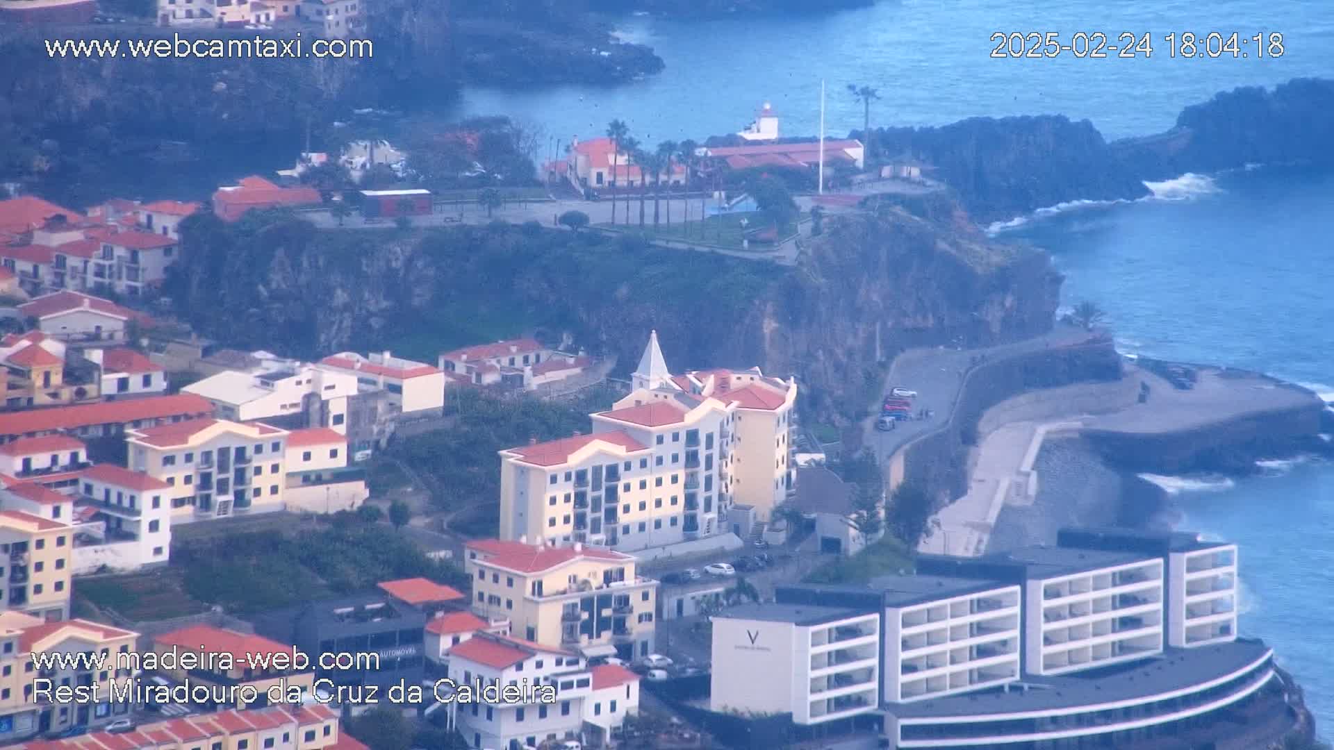 An aerial view shows a coastal town built on a cliffside overlooking a dark blue ocean under an overcast sky.