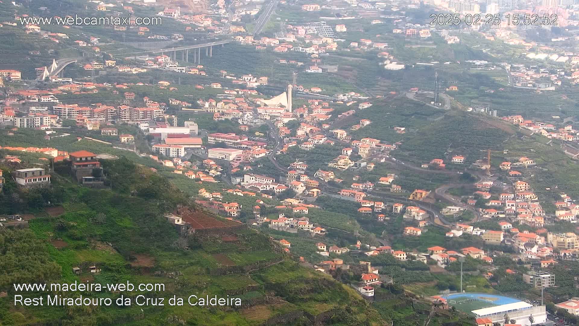 A hazy view of a hillside town with many orange-roofed buildings, terraced farmland, and a bridge in the distance.