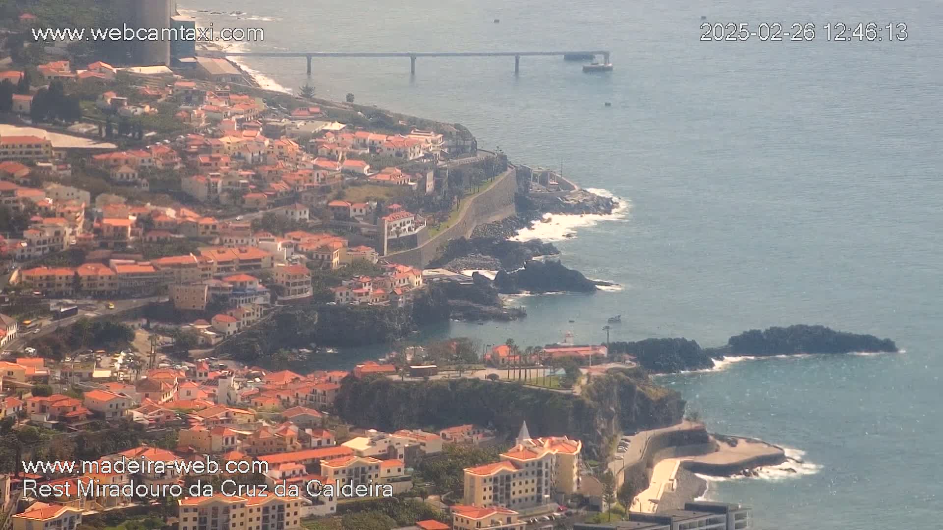 A coastal town with red-roofed buildings clings to a cliffside overlooking a calm ocean with a bridge in the distance.
