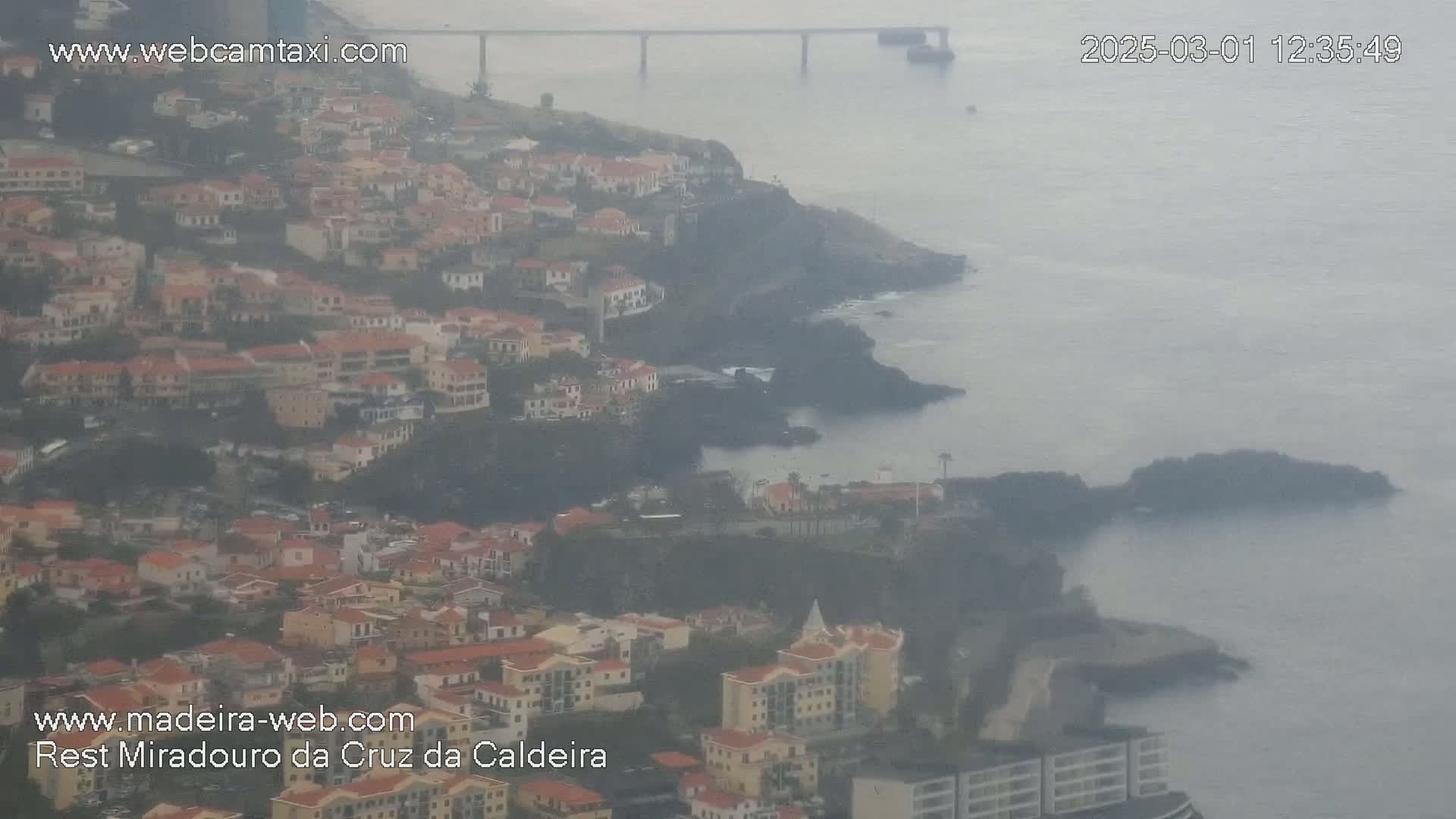 A hazy coastal town with red-roofed buildings clings to a rocky cliffside overlooking a calm ocean and a distant bridge.