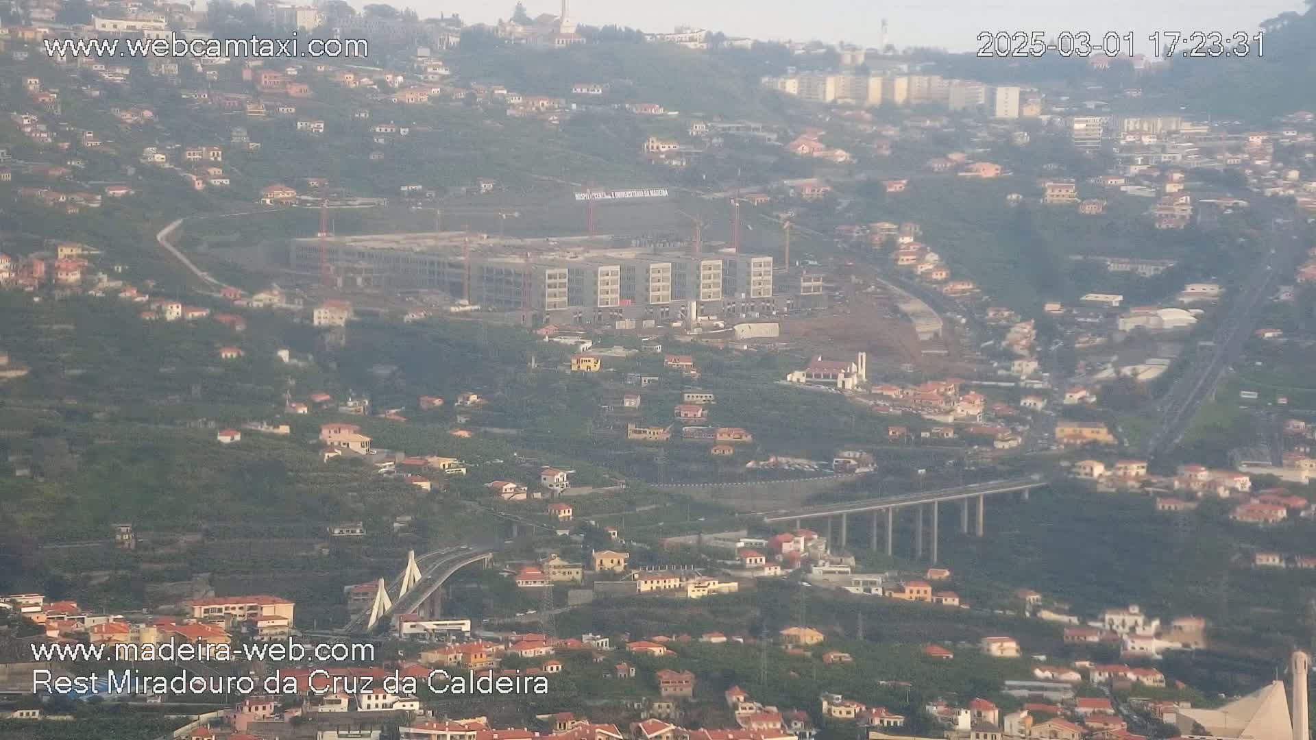 A hazy, high-angle view shows a large building under construction in a hillside town with numerous houses and a long elevated highway and bridge.